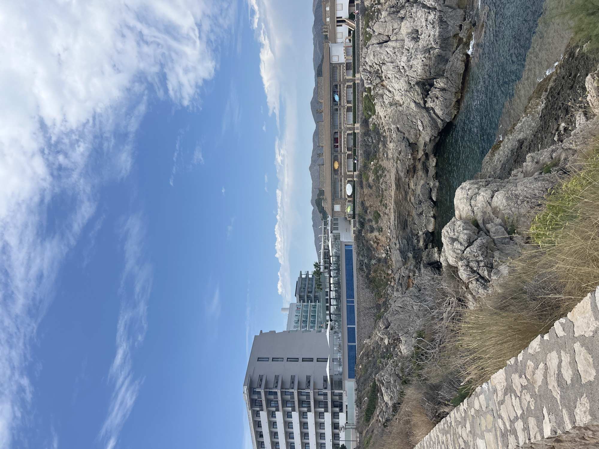 In Cala Lliteres, Spain, a bright, sunny day unfolds with a clear blue sky dotted with wispy white clouds. The scene overlooks a rocky coastline leading to the turquoise waters of the Mediterranean Sea. On the left, a modern, multi-story hotel with a white facade and numerous balconies stands prominently, overlooking a swimming pool area with umbrellas and loungers. Adjacent to it, a lower, more traditional-looking building with arched windows and terracotta roofs stretches horizontally, suggesting a resort or residential complex. The foreground is characterized by a stone pathway and dry grass, with rugged grey rocks leading down to the sea. No people are visible in the frame.