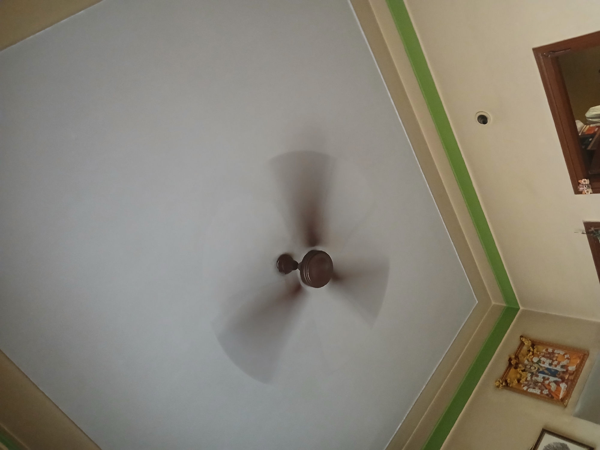 The image is an upward-looking shot of a ceiling in what appears to be an interior space in Bengaluru, India. A ceiling fan with three blades is rotating, causing motion blur. The ceiling is white. Along the edge of the ceiling where it meets the wall, there's a decorative trim with beige and green sections. On the right side of the frame, the wall is painted a light beige color. There are decorative framed artworks hanging on the wall. In the top right corner, a portion of a dark wood-framed mirror or picture is visible. An electrical fixture or sensor is also present on the ceiling near the wall.