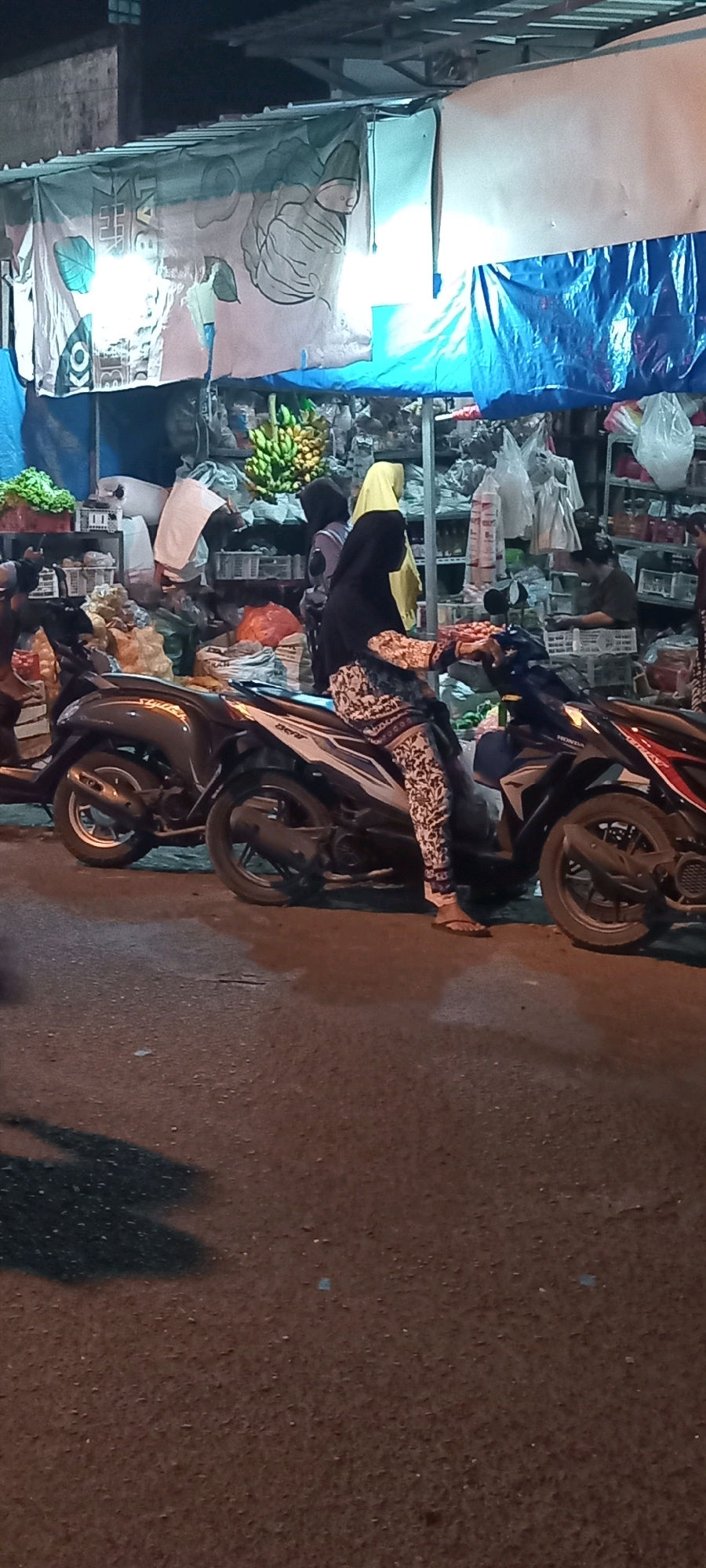 In Babat, Indonesia, a woman wearing a yellow hijab and patterned leggings sits on a dark-colored scooter. The scooter is parked on an asphalt road in front of a market stall. The stall has a blue tarp overhead and displays various goods, including bananas, vegetables, and packaged items. Another woman in a dark hijab is visible behind the first woman, and a third person is seated at a table in the background. Shadows of the scooters are cast on the road.