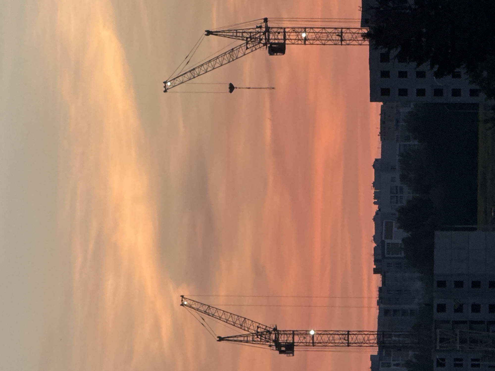 The image shows two large construction cranes silhouetted against a vibrant orange and pink sunset sky. The cranes are positioned on the left and right sides of the frame, with the sky filling the upper two-thirds of the image. Below the cranes, the dark silhouettes of unfinished apartment buildings and trees are visible, suggesting an urban or suburban construction site. The sky has wispy clouds that catch the light of the setting sun, creating a dramatic and colorful backdrop. The scene depicts ongoing construction activity in the city of Palikavichy, Belarus.