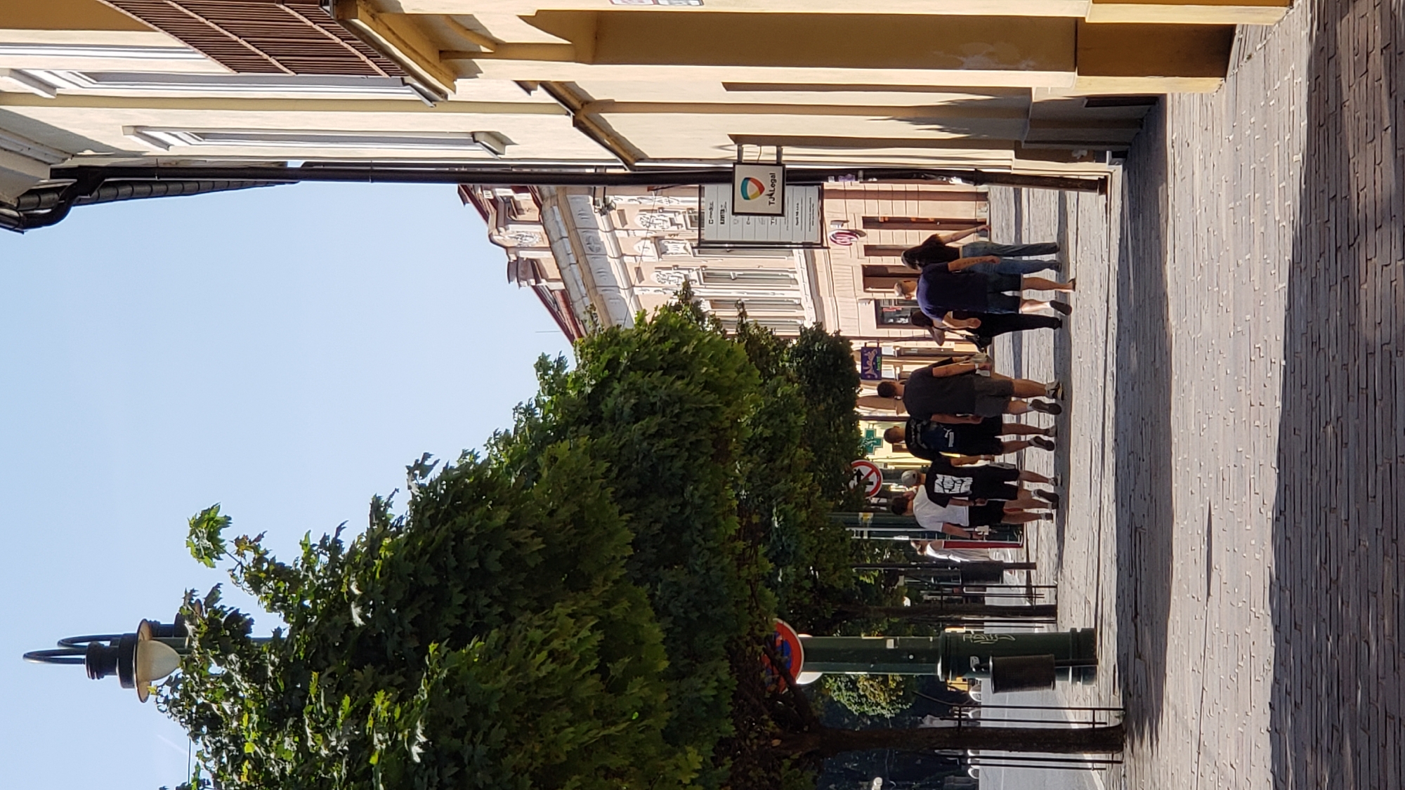 The image depicts a street scene in the city of Prešov, Slovakia, specifically on or near Hlavná Street. A group of individuals, appearing to be a mix of men and women, are walking away from the camera along a cobblestone sidewalk. The people are casually dressed in t-shirts and shorts. On the left side of the sidewalk, a line of trees with full green foliage shades the path. A lamppost with a distinctive curved design stands tall amongst the trees. To the right of the sidewalk, a building with yellow-toned facade and ornate architectural details rises, featuring several windows and a balcony with a wooden railing. Signage is visible on the building, including one that appears to be for a legal service. The sky is clear and blue, suggesting a sunny day. The overall scene conveys a normal urban environment during daylight hours.