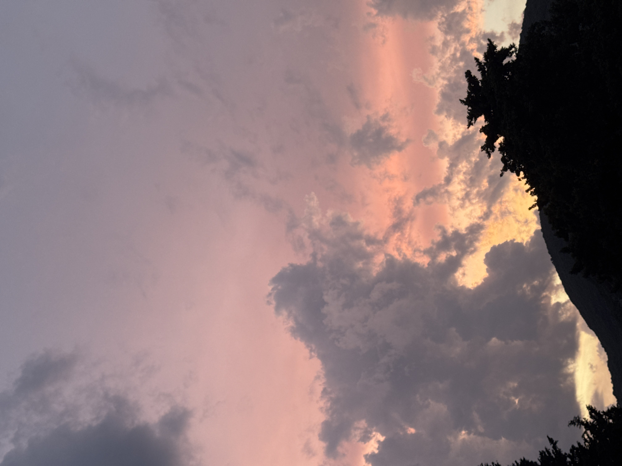 This media file captures a dramatic sunset over a mountainous landscape in Budva, Montenegro. The sky is a canvas of vibrant colors, with soft pinks and oranges blending into cooler purples and grays. Wispy clouds are illuminated by the setting sun, creating a beautiful contrast between light and shadow.
In the foreground, silhouetted trees frame the right side of the image, their dark forms stark against the colorful sky. The rugged outline of a mountain or cliff face is visible on the far right, adding a sense of depth and scale to the scene. The overall atmosphere is one of serene beauty and natural grandeur. The time of day is clearly sunset, as evidenced by the warm, diffused light and the hues in the sky. The weather appears to be clear with scattered clouds, typical of a pleasant evening. The location cues, such as the mountainous terrain and the suggestion of coastal scenery, are consistent with Budva, Montenegro. There is no text visible in the image.