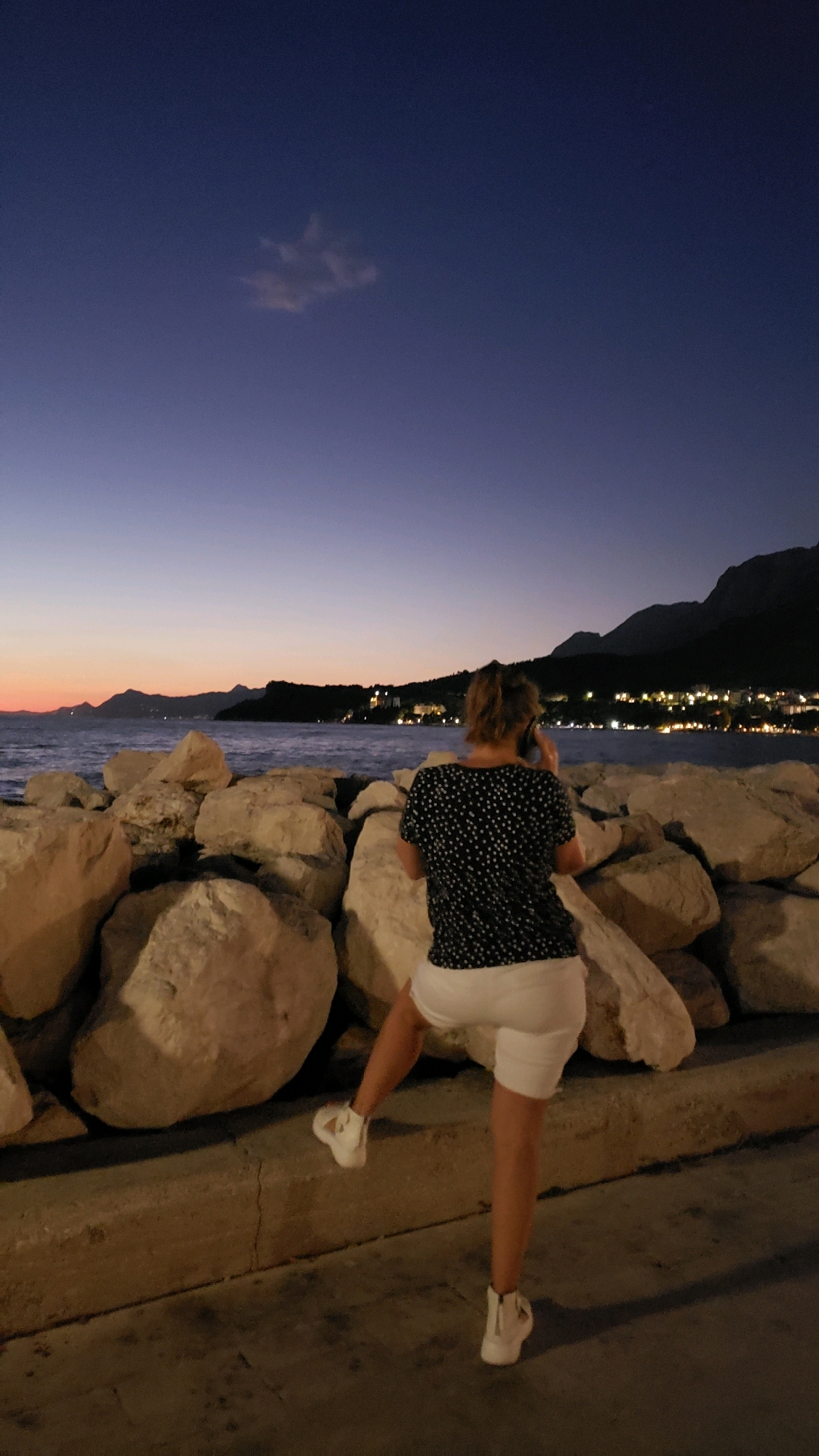 A woman with blonde hair, wearing a dark-colored t-shirt with white polka dots and white shorts, stands facing away from the camera, speaking on her mobile phone. She is positioned on a concrete walkway, with her right leg slightly bent and her left leg extended. The walkway is in front of a barrier constructed from large, light-colored rocks that extend towards the sea. The sea is visible behind the rocks, with waves gently lapping. In the distance, the coastline features hills and mountains, with numerous lights indicating a settlement. The sky above is a deep twilight blue, transitioning to hues of orange and pink near the horizon where the sun has recently set. This scene is located in Općina Tučepi, Croatia, likely along the waterfront promenade.