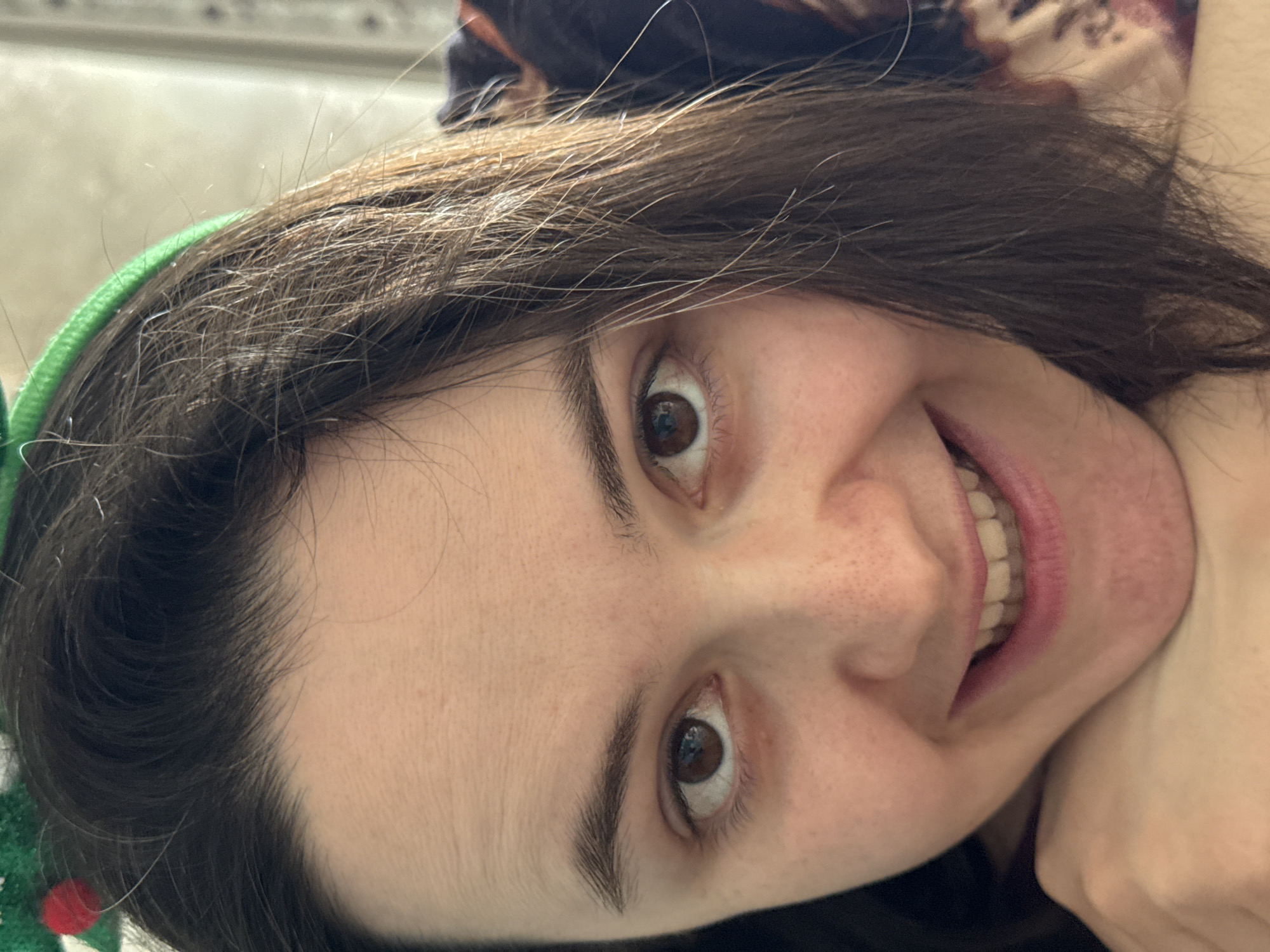 A close-up, slightly rotated image of a woman's face. She has dark brown hair, brown eyes, and fair skin with freckles. She is smiling, revealing her teeth. A green headband is visible in her hair on the left side of the frame. Part of her hand and neck are visible in the lower right corner. The background is out of focus and indistinct. There is no information present in the image that relates to Izhevsk, Russia.