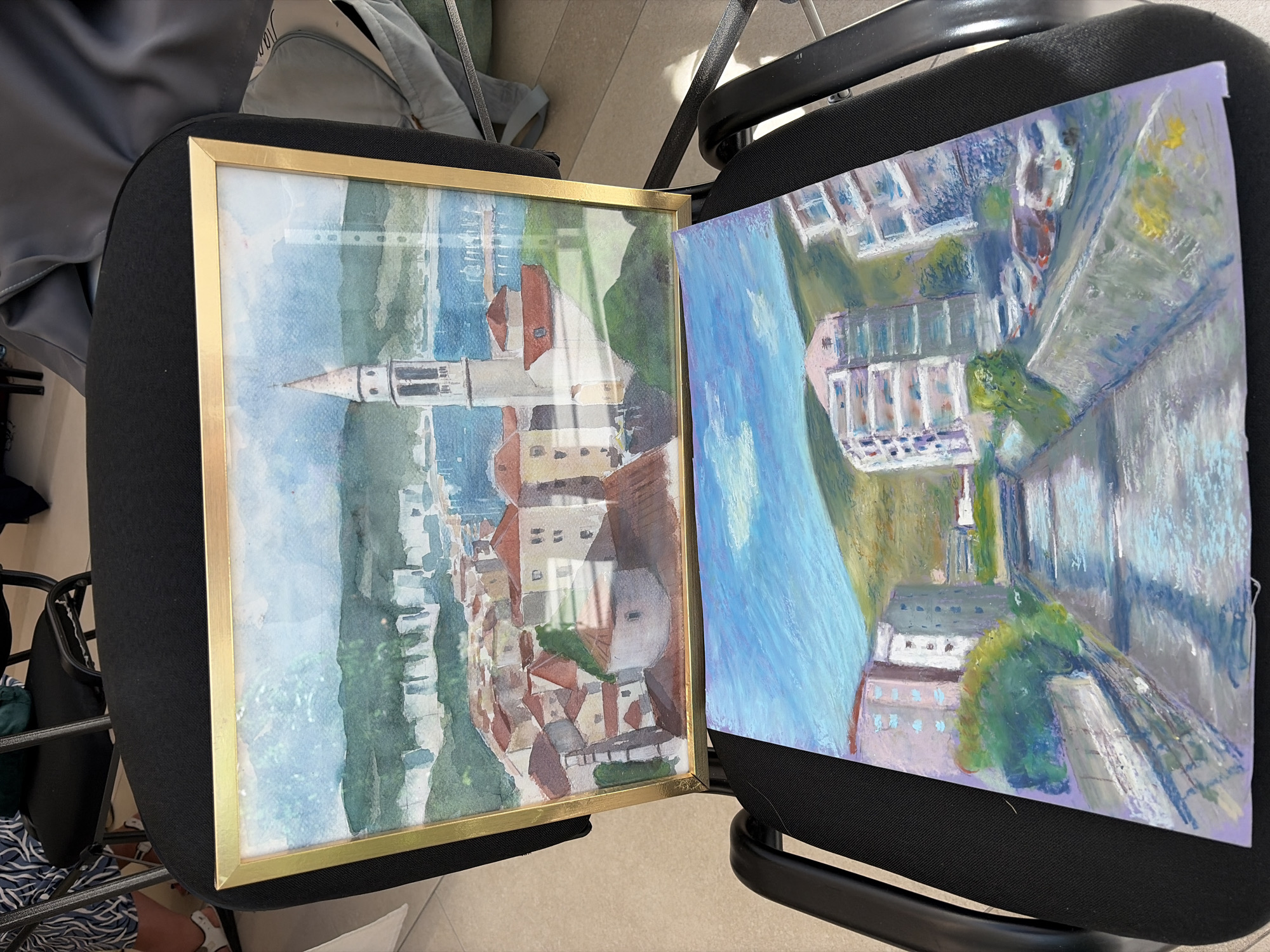 The image displays two paintings placed on a black chair. The top painting, framed in gold, is a watercolor depicting a coastal town with red-tiled roofs and a prominent bell tower, set against a backdrop of green hills and a blue sea with boats. This scene is identifiable as the old town of Budva, Montenegro, with the bell tower of St. John's Church visible. The bottom painting, unframed and rendered in pastel, shows a canal or river lined with modern buildings and a hillside in the background, with cars parked along the side of the canal. The chair is situated indoors, with hints of people and other chairs in the background, suggesting a viewing or exhibition setting.