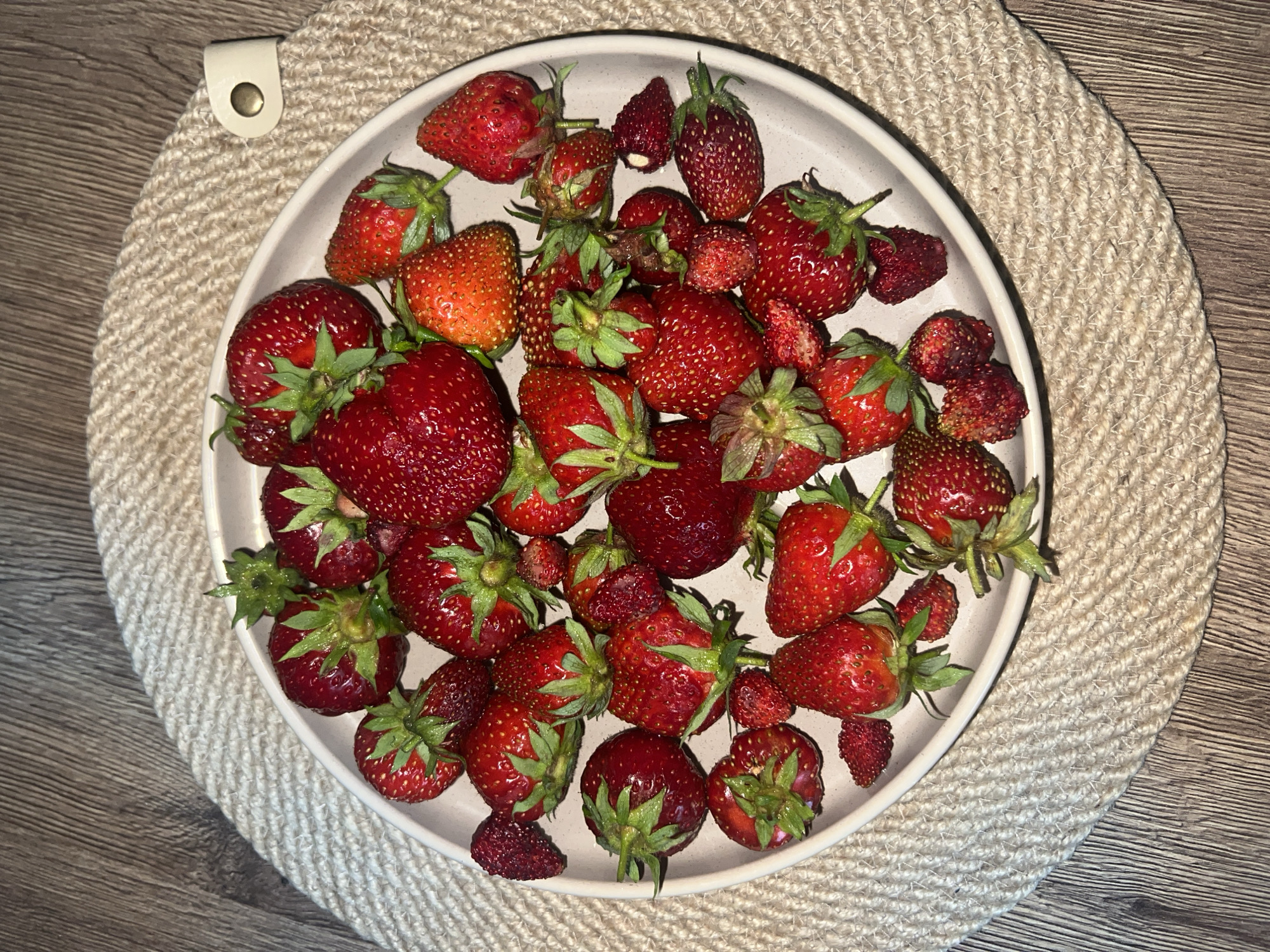 A plate of fresh strawberries sits on a woven placemat on a wooden table. The strawberries are a vibrant red color, with some showing visible seeds and green stems. The plate is round and light-colored. The placemat is made of a coarse, woven material and has a decorative edge. The wooden table surface is visible beneath the placemat and appears to be made of dark wood planks. The image is shot from a top-down perspective. There are no people or animals present in the scene. The lighting suggests an indoor setting. The location is Votkinsk, Russia.