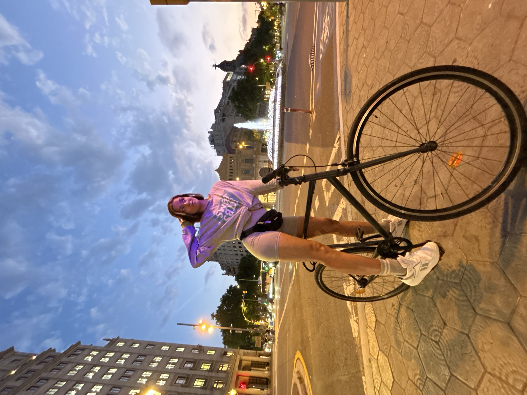 A young woman stands with her bicycle on a tiled sidewalk in Barcelona, Spain, at dusk. She is wearing a light gray long-sleeved sweatshirt, light gray biker shorts, and white sneakers. A black fanny pack is slung across her waist. Her hands are behind her head, and she looks to her left. The bicycle is a dark-colored fixie with a single gear. In the background, a large building with many illuminated windows is on the left. Across the street, a fountain with illuminated water is visible, along with other city buildings, traffic lights, streetlights, and vehicles. The sky is a mix of blue and pink clouds, indicating the transition from day to night. The street is paved with hexagonal tiles.