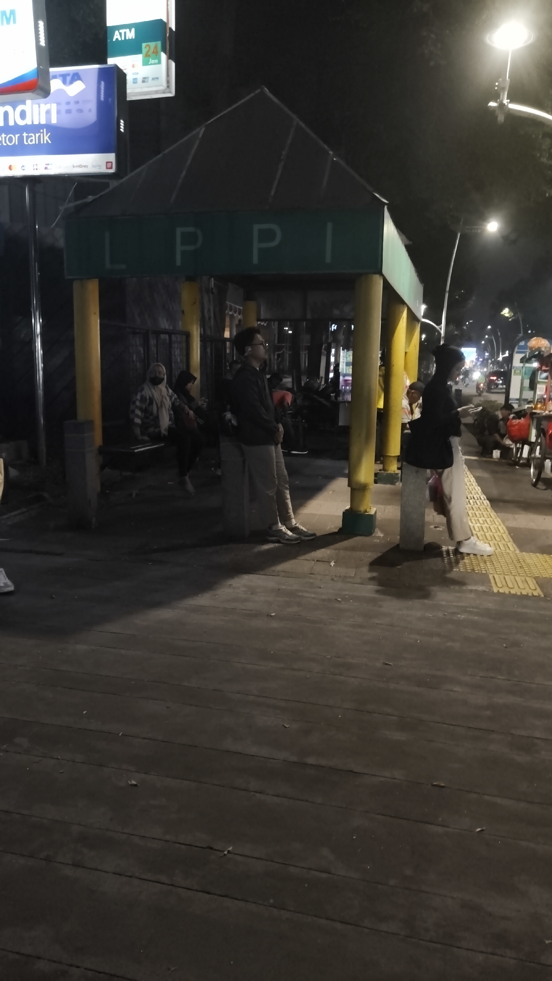 The image captures a nighttime scene at the LPPI bus stop in South Jakarta, Indonesia. A covered shelter with a dark roof and green signage displaying "L P P I" is supported by yellow pillars. Several individuals are present: a man in glasses and a dark jacket stands looking to the right, while another person, also in dark clothing and a head covering, stands looking at their phone. Two women are seated on a bench within the shelter. The ground is paved with dark wooden planks, and a yellow tactile paving strip runs along the edge of the sidewalk. Streetlights illuminate the area, casting shadows. In the background, traffic and other bus stops are visible, along with neon signs and parked vehicles, suggesting a busy urban environment.
