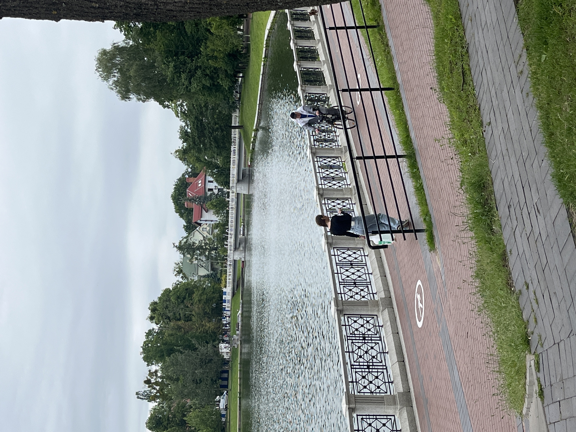 The image depicts a scenic view of a park with a lake in Kaliningrad, Russia. In the foreground, a paved walkway is visible, made of reddish-brown bricks interspersed with dark gray stone slabs. A section of this walkway features a circular white symbol with a line through it, possibly indicating a prohibition. To the left of this walkway, a woman wearing a black top and blue jeans is walking, carrying a white plastic bag. To the right, a person on a bicycle, wearing a grey hooded jacket and white pants, is riding along the walkway.
The walkway is bordered by a black metal fence with ornate lattice work. Beyond the fence lies the lake, its surface rippling with the wind. The lake is surrounded by a grassy bank and a line of trees. Across the lake, several buildings with red and green roofs are visible, nestled among the trees. A white pedestrian bridge spans a portion of the lake in the background. The sky is overcast and cloudy, suggesting a cool or temperate day. The overall scene conveys a sense of peaceful recreation in an urban park setting.