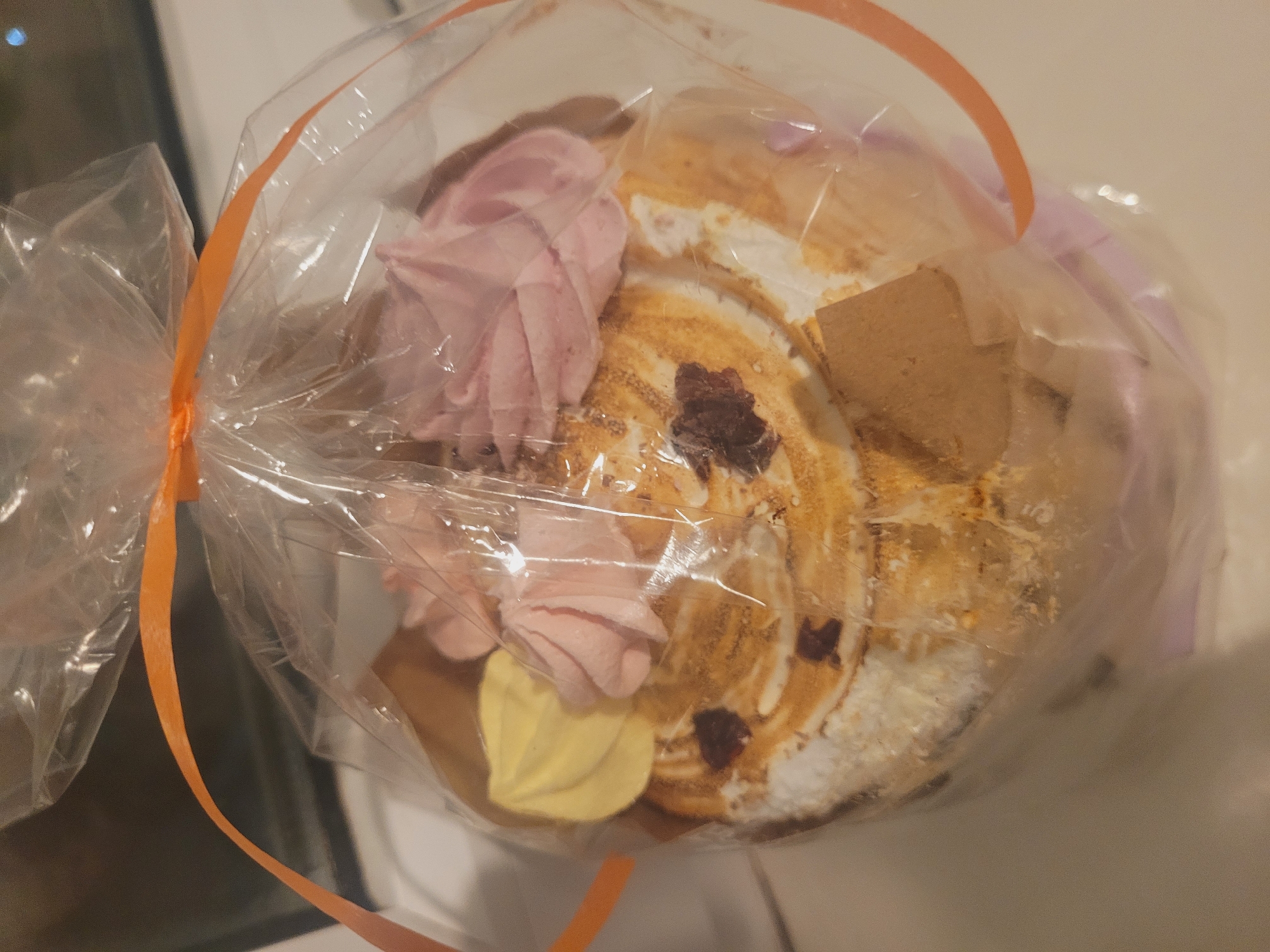 A packaged dessert sits on a light-colored surface. The dessert is encased in clear plastic, tied with an orange ribbon. The dessert features light pink and yellow frosting rosettes. The body of the dessert appears to be a meringue-like swirl with browned spots and scattered dark pieces. A square of what appears to be shortbread is visible on the dessert's surface. The image does not contain information regarding the location of the city of Izhevsk, Russia.