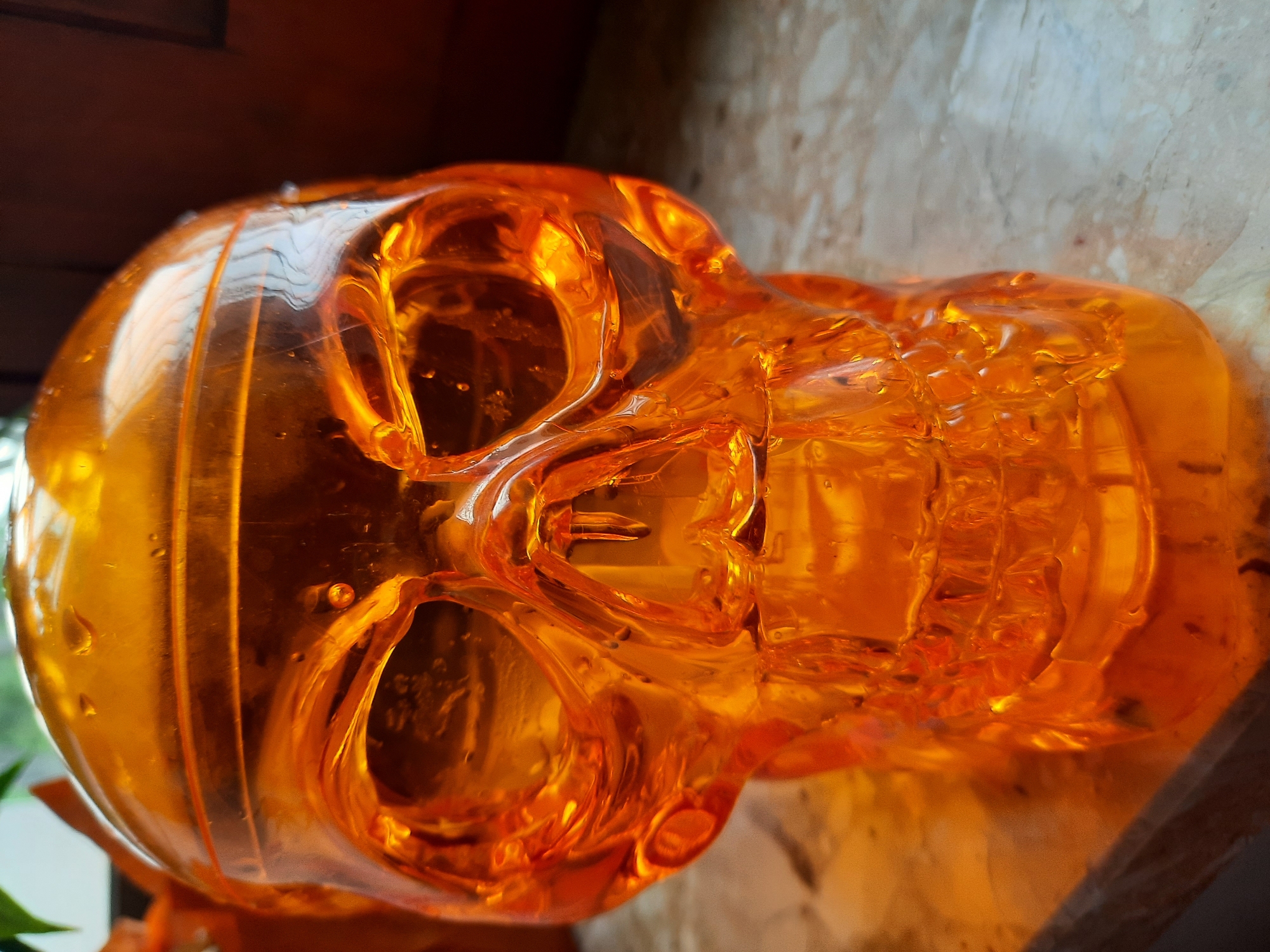 The image shows a close-up, angled view of an orange, transparent skull-shaped object. The object appears to be made of plastic or glass and has a glossy finish. There are small bubbles and imperfections visible within the material, suggesting it might be a decorative item or a container. The skull is positioned on a marble-like surface with a subtle texture. In the background, a blurred area of warm tones suggests wooden furniture or a wall. The lighting creates highlights and shadows, giving the object a somewhat translucent and eerie appearance. There is no indication of the city of Rosenheim, Germany, in the image.