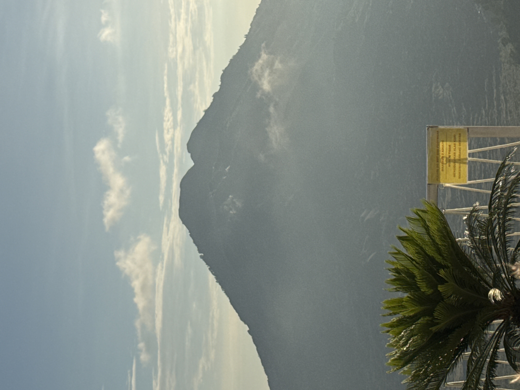 The image shows a tall, imposing mountain looming in the background. The sky is blue with scattered clouds. In the foreground, a tropical plant, possibly a palm or cycad, partially obscures a sign written in both English and Indonesian. The sign reads, "PLEASE DO NOT WALK FOOTWEAR MAKSIMUM 2 ADULTS MOHON TIDAK MENGGUNAKAN ALAS KAKI MAKSIMAL 2 ORANG DEWASA" indicating a request to remove footwear, with a maximum of two adults allowed. The location is likely Kedisan, Indonesia, based on the Indonesian language on the sign and the tropical vegetation. The scene appears to be a scenic overlook or viewing point near the mountain.