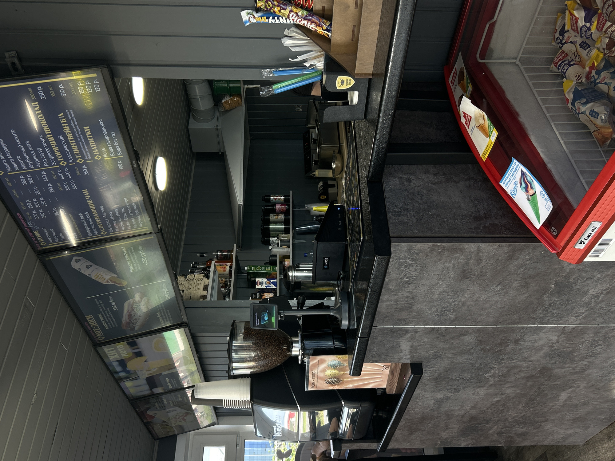 The image captures an indoor view of a cafe or food kiosk, featuring a service counter with various commercial equipment and extensive menu boards. The scene is well-lit by interior lights, with some warm-toned, oval-shaped lights visible on the ceiling.
In the foreground and midground, a dark counter stretches across the frame. On it, a professional coffee machine with a grinder full of coffee beans is prominent. Next to it is a digital display showing "MEIN NELLI" and "Dere is no internet connection". Various bottles, possibly syrups or flavorings, are neatly arranged on shelves behind the counter. A dispenser holds napkins and blue and green plastic straws. Nearby, a brown paper bag contains small snack items, including a "Picnic" branded bar. A display for "Кейк-попс" (Cake Pops) with a price of "180" is also visible on the counter.
To the left, several large, illuminated menu boards are mounted on a grey, paneled wall. These boards are in Russian and list a wide array of beverages and hot dogs. Categories include "КОФЕ" (Coffee) with items like Americano, Cappuccino, Latte, and Raf, along with prices ranging from 240 p. to 440 p. "СОГРЕВАЮЩИЕ ЧАИ" (Warming Teas) like Sea Buckthorn Raspberry and Kiwi-Basil are listed, priced around 200 p. to 250 p. "ГОРЯЧИЙ ШОКОЛАД" (Hot Chocolate) and "ГЛИНТВЕЙН Б/А" (Non-alcoholic Mulled Wine) are also on offer. A section for "НАПИТКИ" (Drinks) lists Cola, sparkling, and still water, with prices from 100 p. to 150 p. Another section advertises "ХИТ Хот-доги" (HIT Hot Dogs) including "Нордик чикен" (Nordic Chicken) at 376 rub/dog, "Нордик биф" (Nordic Beef) at 396 rub, and "Нордик классика" (Nordic Classic) at 250 rub/dog.
To the far right, a red-rimmed commercial freezer or cooler is visible, filled with various packaged ice cream products. Labels on the ice cream include "Корнето", "КЛУБНИКА БАБЛ-ГАН" (Strawberry Bubble Gum), and "ЧЕРНАЯ СМОРОДИНА" (Black Currant). The floor is a dark grey tiled surface. No people are present, indicating a paused moment in what appears to be a bustling service area.