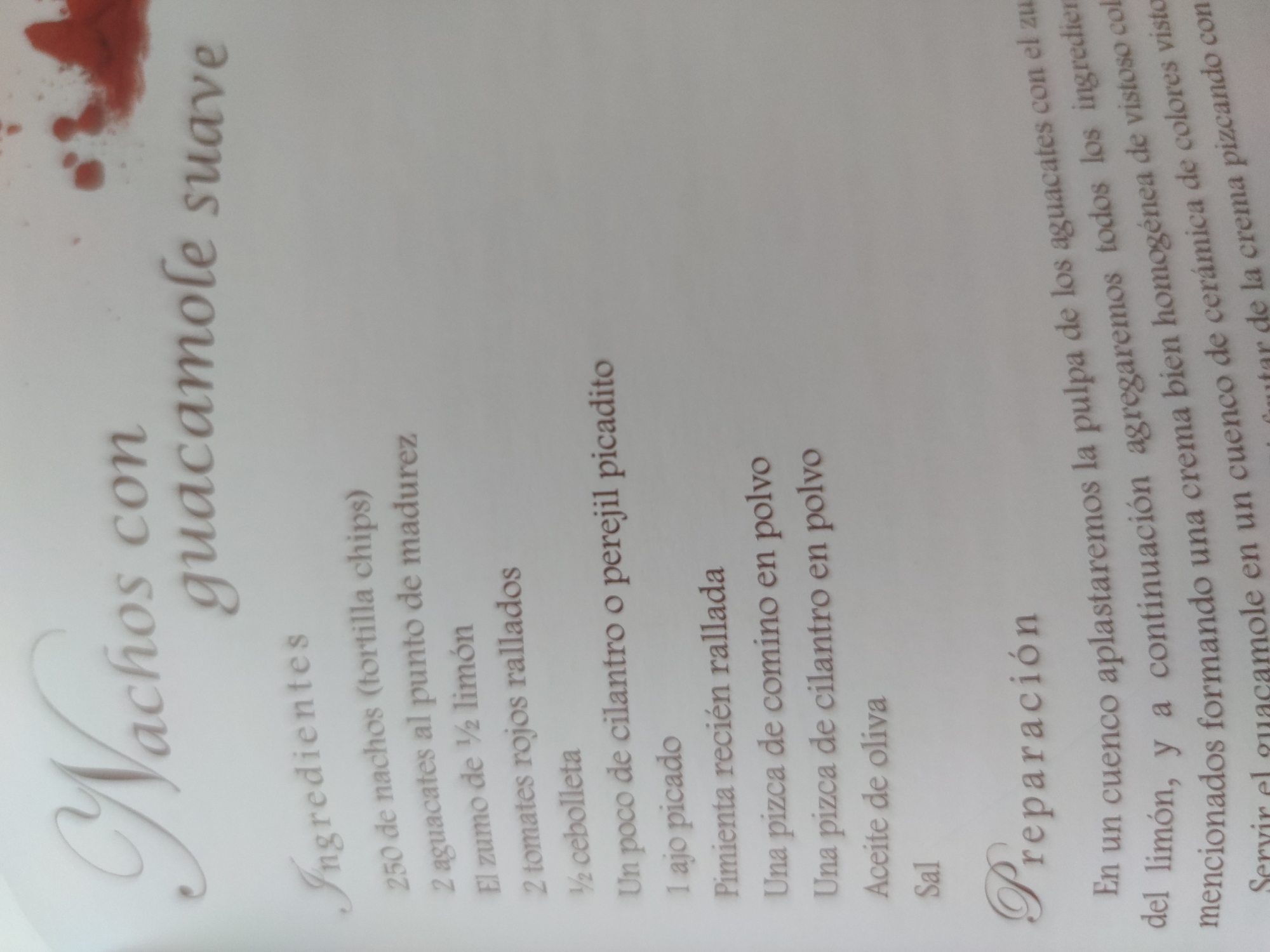 Here is a detailed description of the image:
The image is a close-up shot of a recipe for "Nachos con guacamole suave" (Nachos with smooth guacamole). The text is printed in Spanish. The image is slightly blurry and rotated.
The recipe lists the ingredients:
- 250 nachos (tortilla chips)
- 2 avocados at the point of ripeness
- Juice of 1/2 lemon
- 2 grated red tomatoes
- 1/2 onion
- A little chopped cilantro or parsley
- 1 chopped garlic clove
- Freshly grated pepper
- A pinch of cumin powder
- A pinch of cilantro powder
- Olive oil
- Salt
The recipe instructions, under the heading "Preparación" (Preparation), advise mashing the avocado pulp in a bowl with the lemon juice, then adding all the ingredients and mixing well to form a homogeneous cream with a vibrant color. The guacamole should be served in a ceramic bowl of vibrant colors and enjoyed by scooping it with tortilla chips.