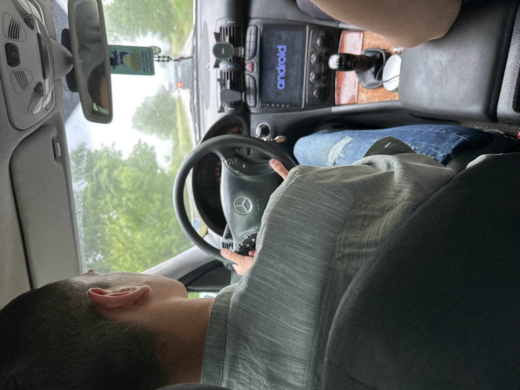 A person is driving a car. The dashboard shows an Android system. The vehicle is on a road with green trees visible through the windshield. The driver's left hand is on the steering wheel. The vehicle appears to be a Mercedes-Benz, based on the steering wheel logo. The location is not verifiable as Novo Selo, Bulgaria, based on the image alone.