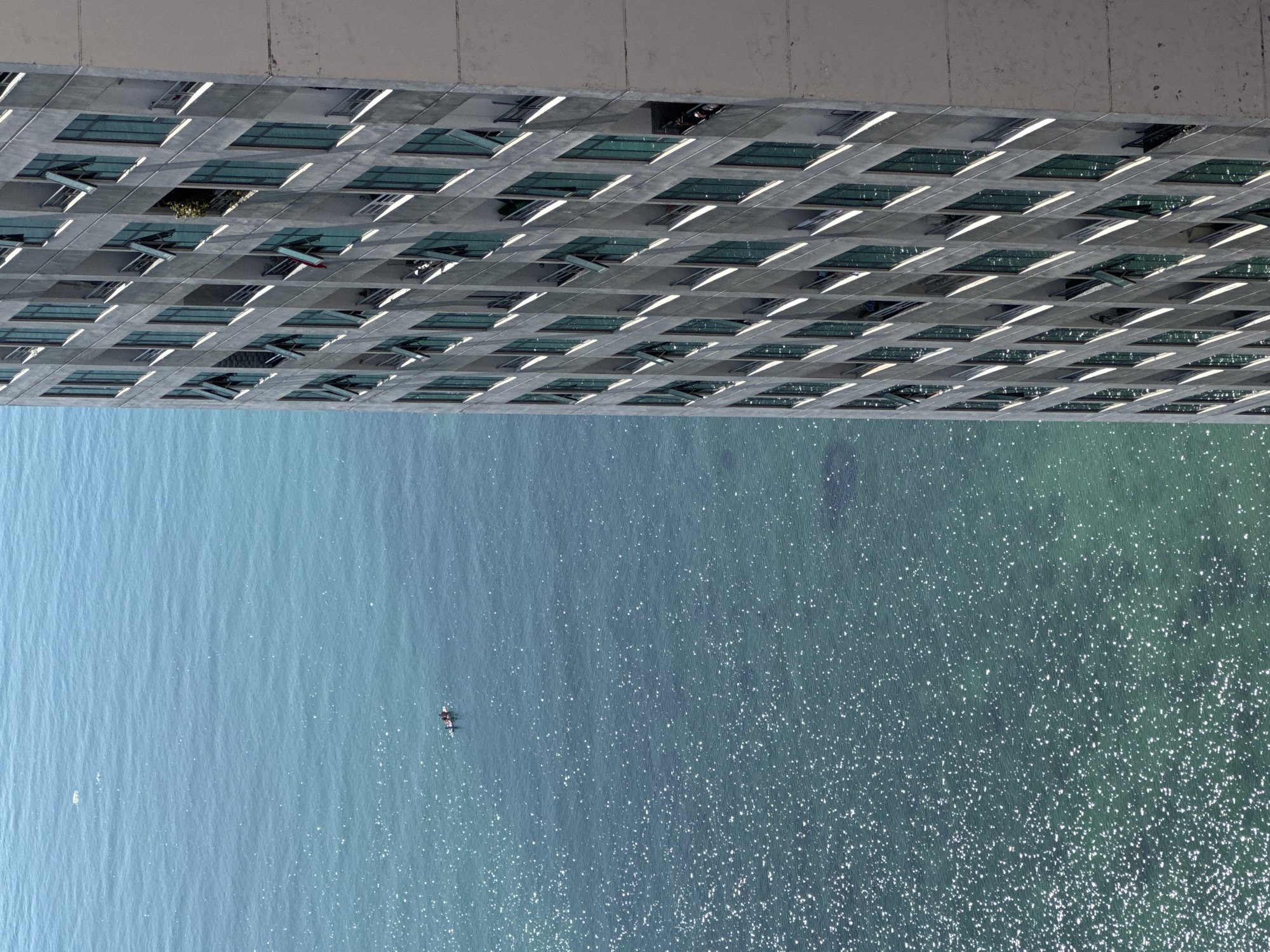 A high-angle, medium shot captures a portion of a modern, multi-story building adjacent to a body of shimmering turquoise water. The building, rendered in shades of gray, features a repetitive pattern of windows, many of which are open, revealing glimpses of balconies and interior spaces. Some balconies have plants, and a red flag is visible from one window. In the water, a small boat with two occupants is visible, contributing a sense of scale and human presence against the vastness of the ocean. The water's surface is marked by numerous bright, sunlit reflections, creating a dappled effect. The overall scene suggests a coastal urban environment in Bac Nha Trang Ward, Vietnam, with a juxtaposition of man-made architecture and natural scenery.