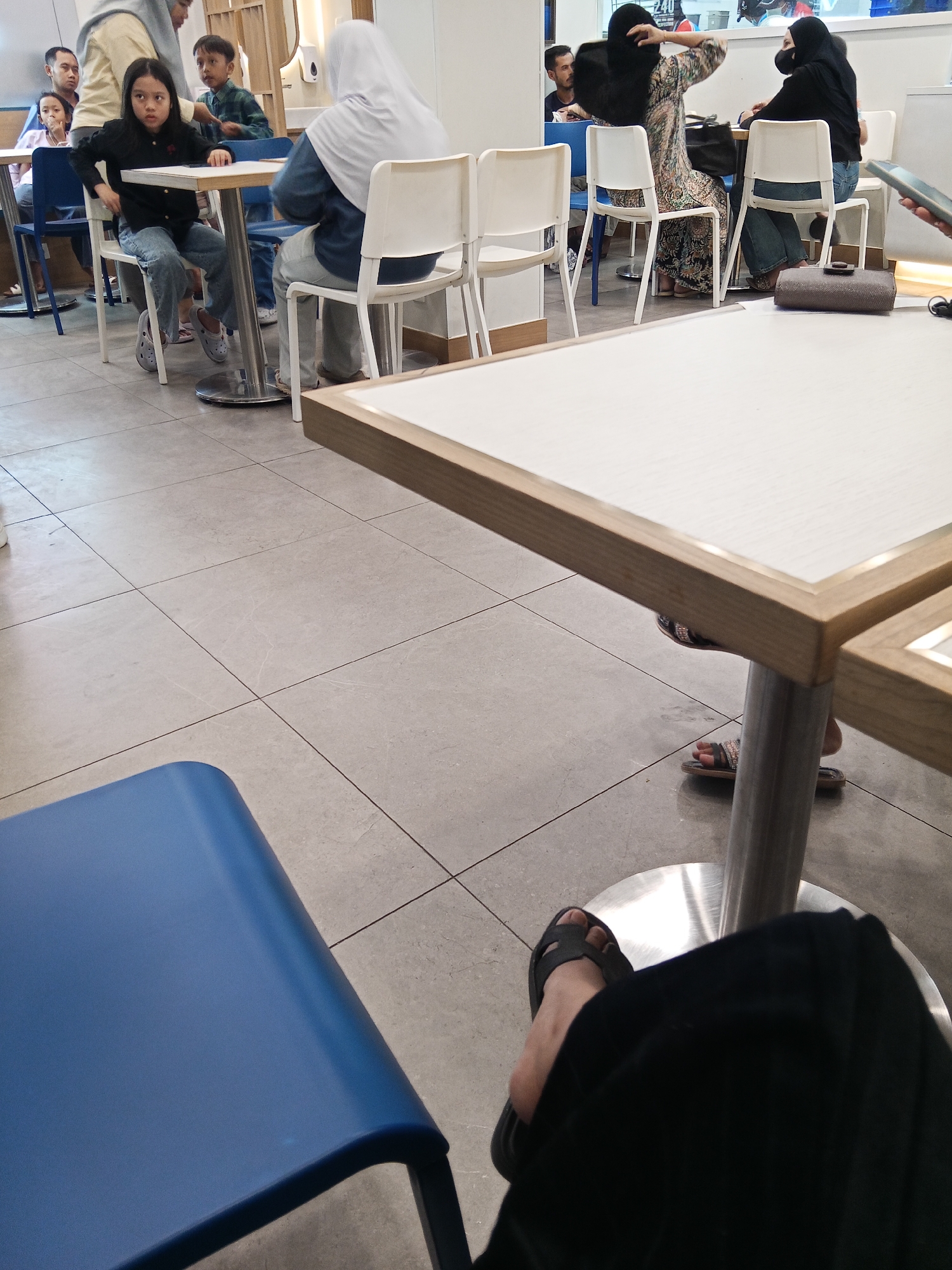 The image depicts a group of people seated at tables inside a building, likely a restaurant or food court, with tiled flooring. In the foreground, a person's legs and feet are visible, with one foot wearing a black sandal. A blue chair is also partially visible. In the mid-ground, a young girl with long dark hair sits at a table, wearing a black shirt and blue jeans. To her left, a man and a younger boy are seated. Across from them, a person wearing a white hijab and a blue top is seated. Further back, two women wearing hijabs are seated at another table; one in a patterned dress and the other in a black top and jeans. The environment suggests an indoor dining area with multiple tables and chairs. The scene is consistent with a public dining space, such as a food court or cafe in Lubuklinggau, Indonesia.