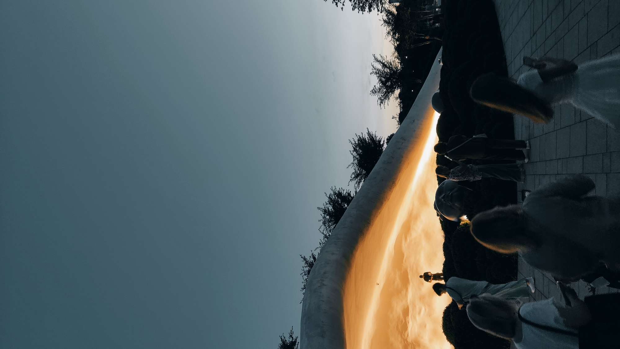 The image depicts a group of people gathered near a large, curved, reflective sculpture. The sculpture is catching the warm, golden light of what appears to be a sunset or sunrise, casting a bright orange glow on its surface. The sky above is a deep, dusky blue, suggesting the time of day is either early morning or late evening. Trees with dark foliage are visible behind the sculpture and to the side, framing the scene. The people are standing on a paved area, possibly a plaza or park, and their silhouettes are cast against the illuminated sculpture and the darker background. Some individuals appear to be interacting or observing the sculpture, while others are walking or standing. One person in the foreground on the right seems to be holding a phone, possibly taking a photograph. The overall mood is serene, with the dramatic lighting from the sculpture creating a visually striking effect. There is no visible text in the image. The location context provided is Krasnodar, Russia, which might imply an outdoor public space within the city.