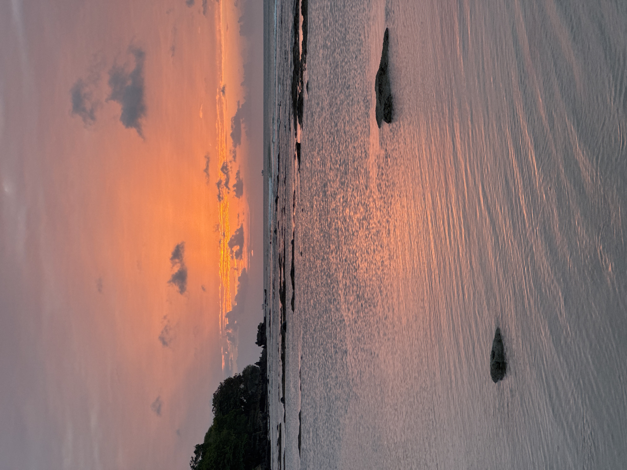 This is a vertical, eye-level, medium shot of a sunset over the ocean in Kuta Selatan, Indonesia. The sky is a gradient of colors from a soft pink and lavender at the top to a vibrant orange and yellow near the horizon. The clouds are scattered and wispy, some tinged with pink from the sunset. In the distance, a ship is visible on the horizon. The ocean is calm with gentle ripples, reflecting the colors of the sky. In the foreground, a few dark rocks are partially submerged in the water, breaking the surface. To the left, a lush green hill with trees slopes down to the rocky shoreline. The overall mood is serene and peaceful.