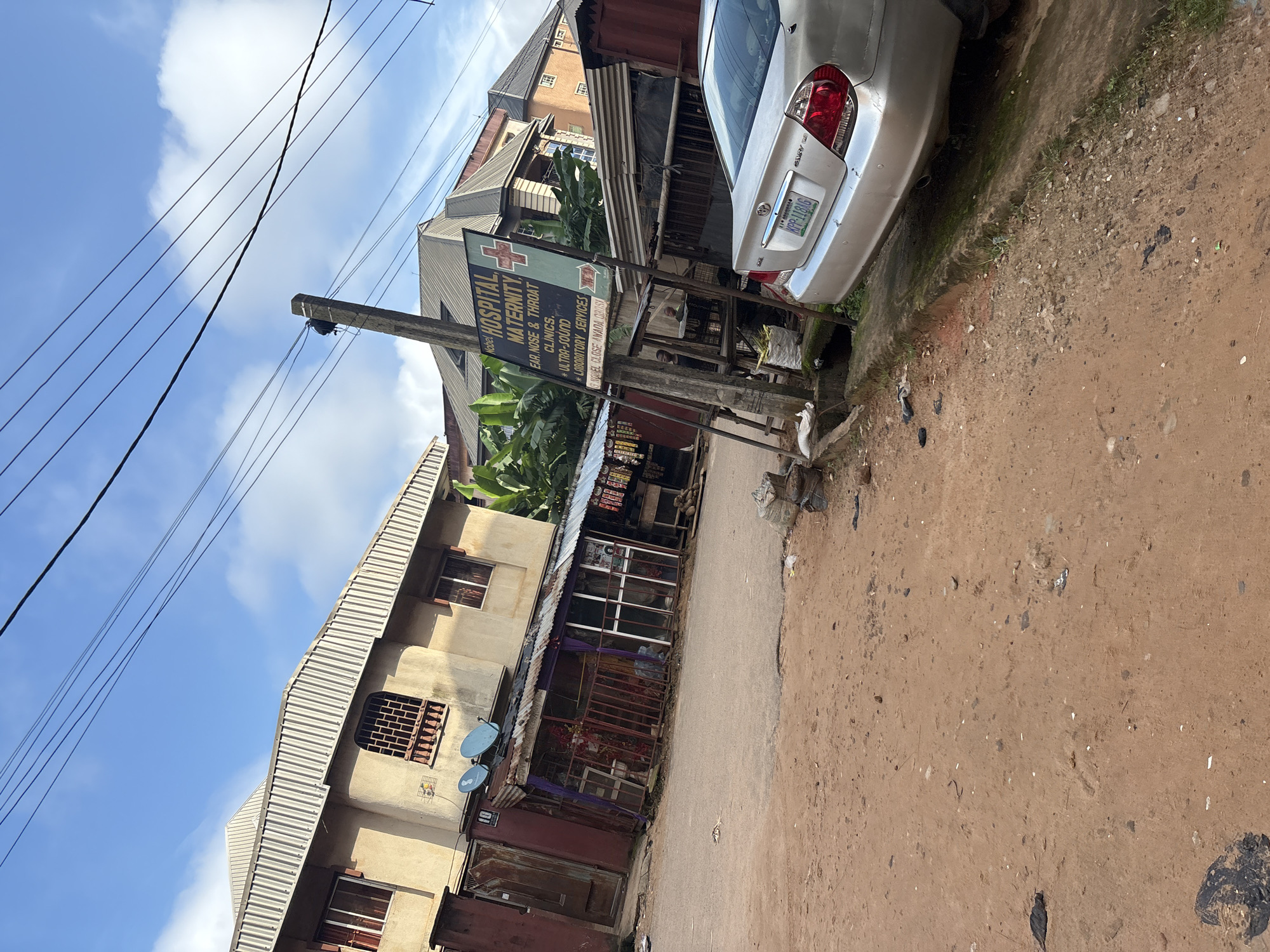 The image shows a street scene in Onitsha, Nigeria. A silver Toyota Camry is parked on the right side of the image, facing away from the viewer. A signpost for "Nobel Hospital Maternity" is visible in the center of the image, listing services such as "Ear, Nose & Throat Clinics", "Ultra-Sound", and "Laboratory Services." Adjacent to the hospital sign is a small stall with various items displayed. In the background, buildings with corrugated iron roofs are visible. The street appears to be unpaved with some debris scattered on the ground. The sky is blue with scattered clouds. There are two satellite dishes on the roof of a building on the left. The overall impression is of a bustling urban environment with a mix of residential and commercial structures.