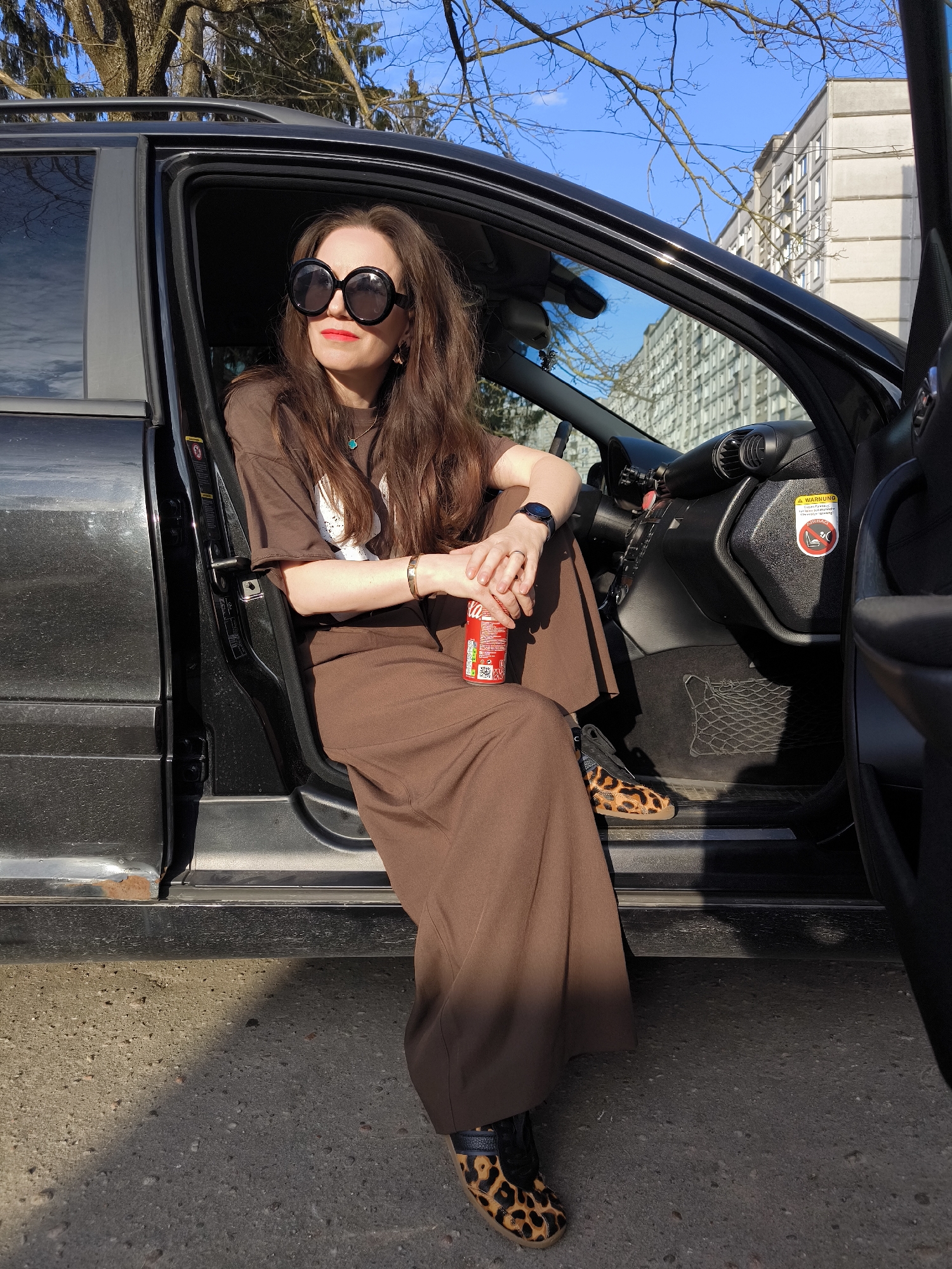 A medium shot captures a woman seated in the driver's seat of a black car with the door open. The woman has long brown hair, is wearing large round sunglasses, and red lipstick. She is dressed in a brown outfit. She is holding a can of Coca-Cola and wearing leopard print shoes.
The car is parked on an asphalt surface that shows signs of wear and tear, including small cracks and patches. A large, multi-story building is visible in the background, suggesting an urban location. Based on the architectural style and surrounding environment, it is possible that this photo was taken in Riga, Latvia. The sky is partially visible with bare branches. The lighting suggests it is daytime, with strong shadows cast on the asphalt.