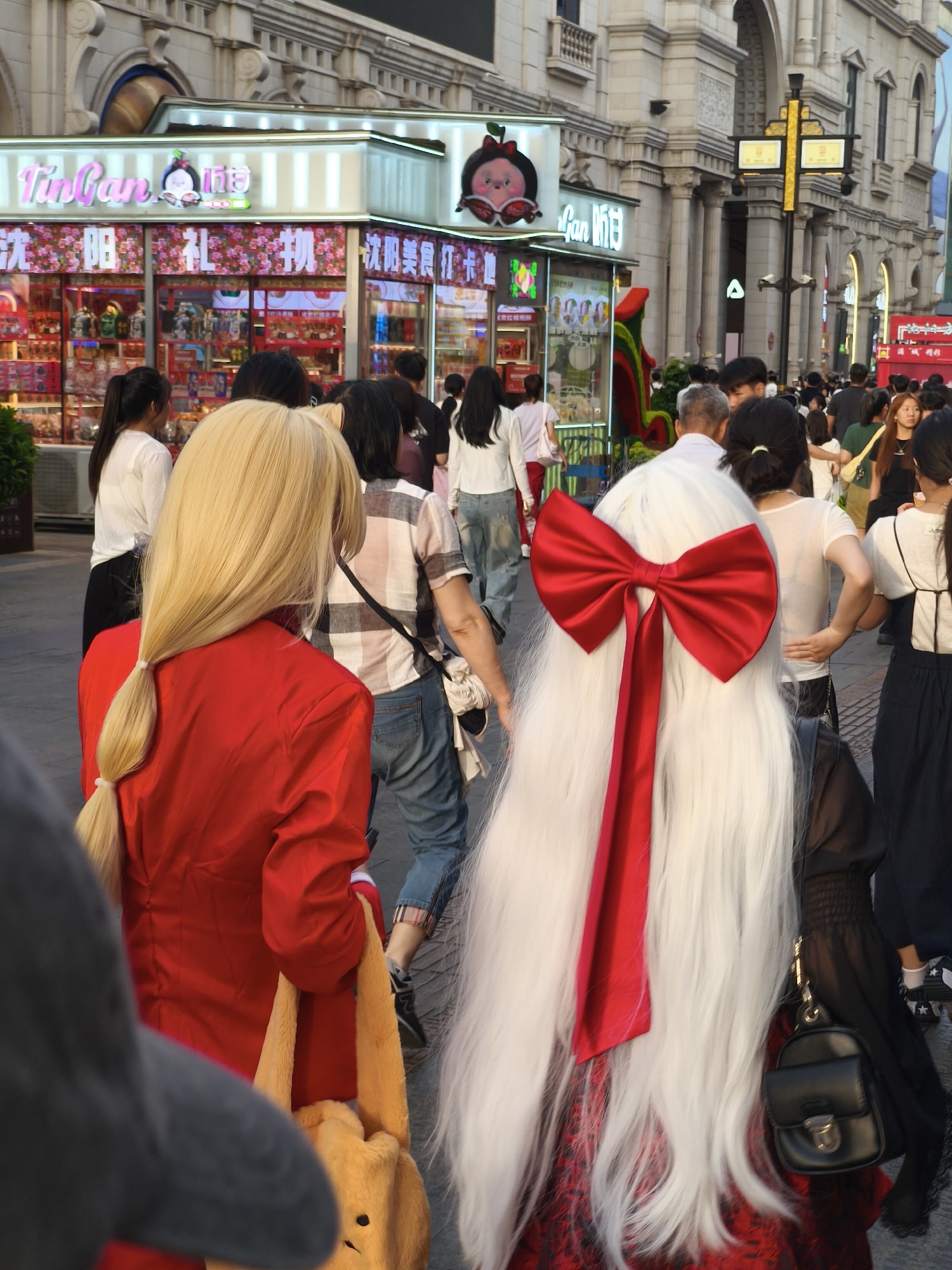 A street scene in Shenhe District, China, features a bustling pedestrian area with shops lining the street. In the foreground, two individuals are prominent. One person, seen from the back, has long, blonde hair tied in a ponytail and is wearing a red jacket and a yellow plush bag. To their right, another person is styled with a voluminous white wig adorned with a large red bow. Several other people are visible in the midground and background, walking and browsing. The shops display various goods, including what appear to be novelty items and food. The architecture suggests a commercial district with decorative building facades and street lighting. The overall atmosphere is lively and crowded, indicative of a popular urban shopping area.