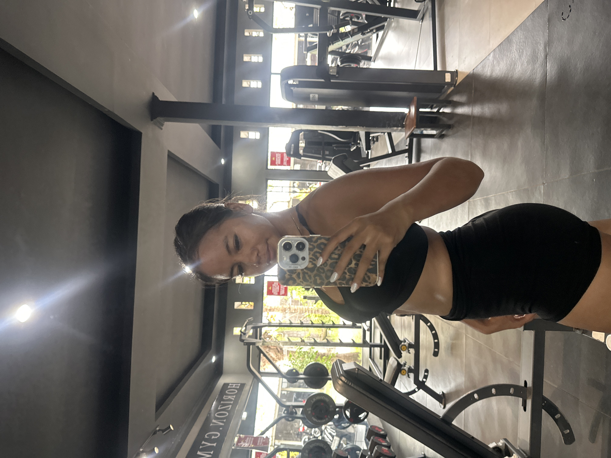 A woman is observed taking a mirror selfie inside Horizon Gym in Batubulan Kangin, Indonesia. She is positioned facing a large mirror, holding a smartphone with a leopard print case in her right hand. The woman has dark hair and is wearing a black sports bra and black shorts. Her fingernails are painted white. The background of the image displays the gym's interior, featuring various exercise equipment including weight benches with yellow accents, dumbbell racks with weights, a squat rack, and other strength training machines. Large windows behind the equipment offer a view of an outdoor environment with trees and foliage. The gym floor is composed of dark grey and lighter beige square tiles, while the ceiling is dark grey with several recessed lights. The reversed text "HORIZON GYM" is visible on the mirror's surface.