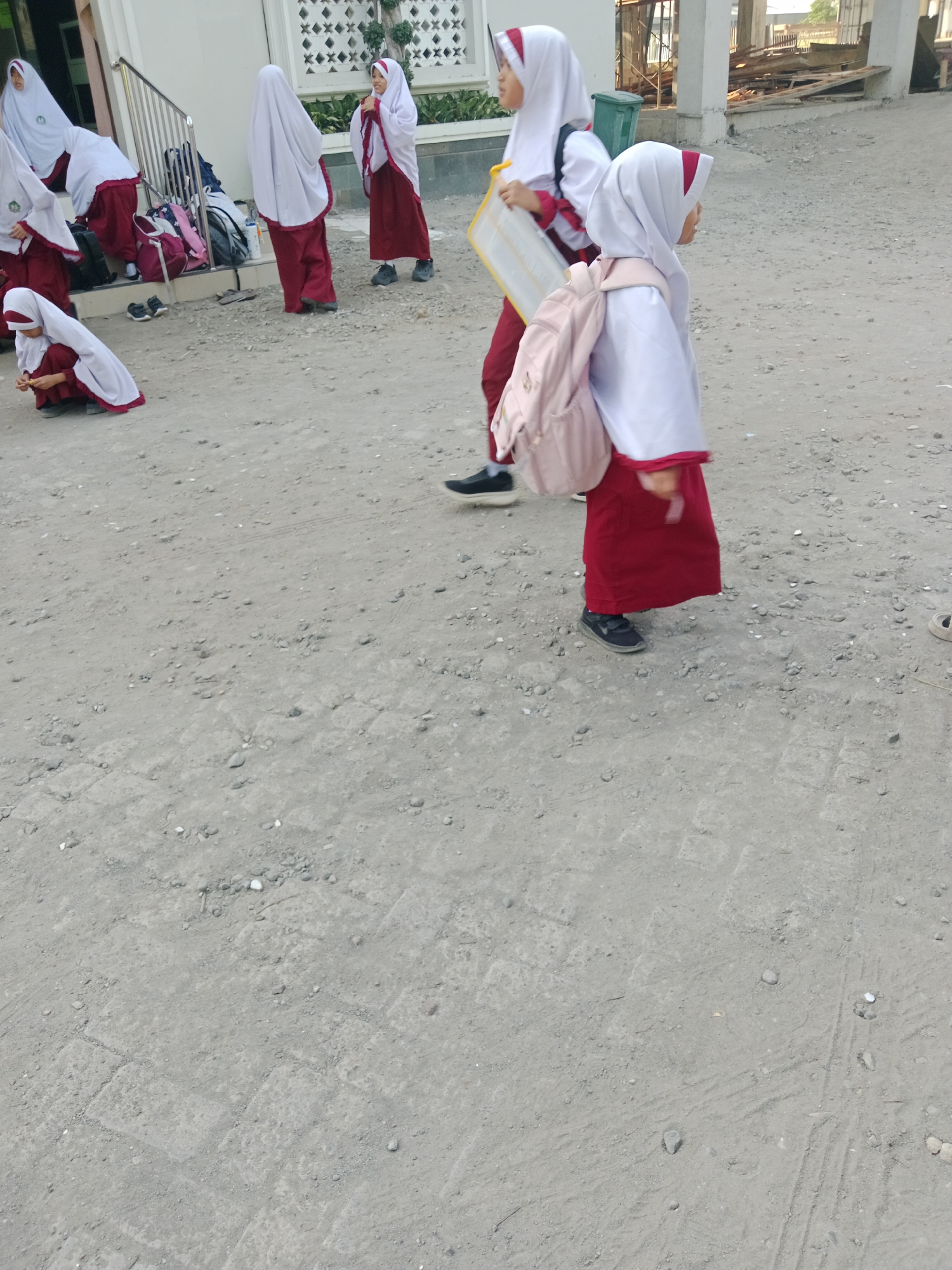A group of students wearing traditional Indonesian school uniforms, consisting of white hijabs and red skirts, are gathered on a dirt path. The students are of various ages and are engaged in different activities. Some are standing and talking, while others are sitting or walking. One student is holding a yellow board. Backpacks and other school items are scattered around them. The background shows a building with white walls and a fence. The scene appears to be an outdoor area, possibly a schoolyard, in Kediri City, Indonesia. The lighting suggests it is daytime.