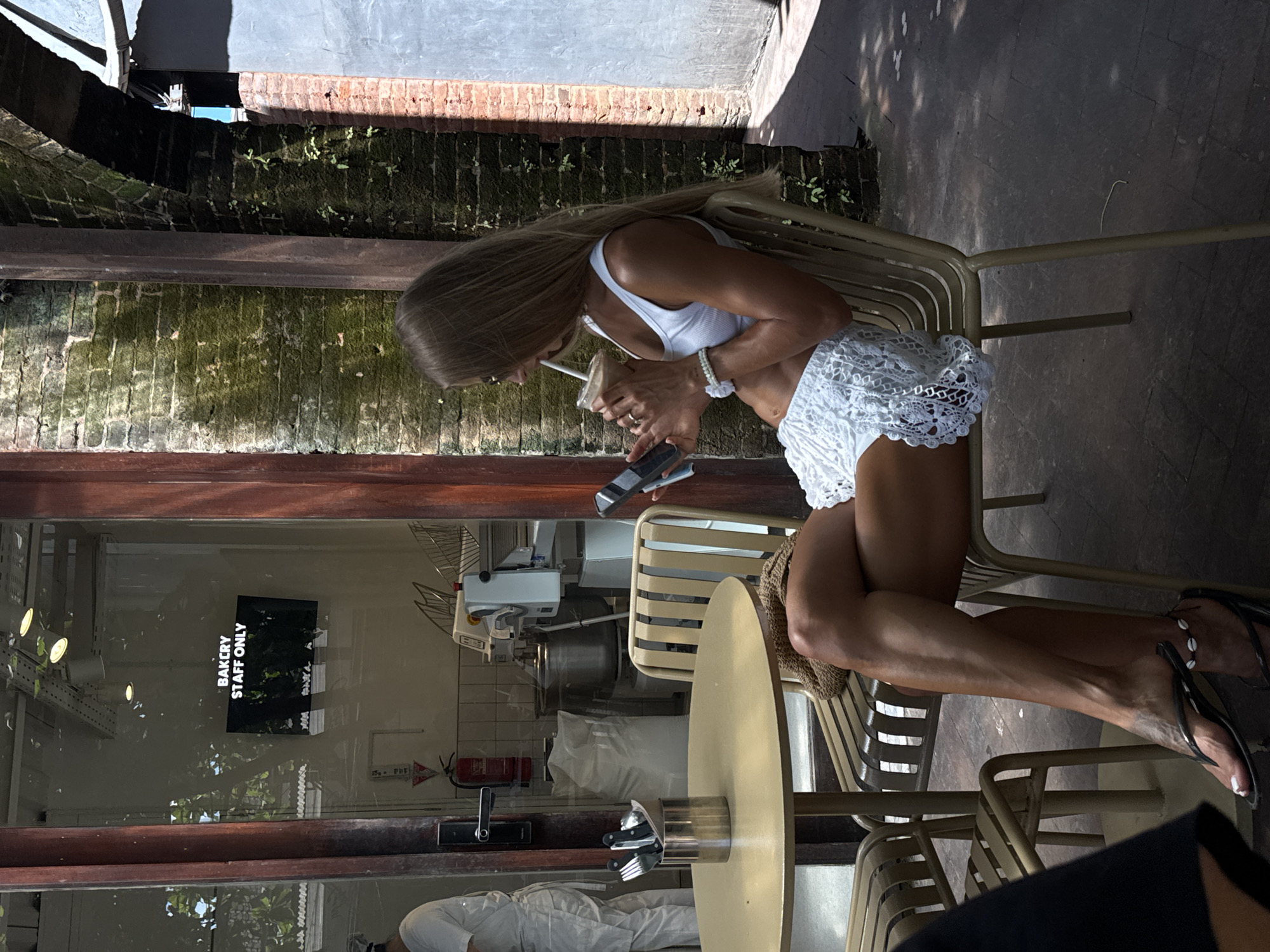 A tanned woman with long blonde hair is seated in a metal chair outdoors, drinking from a clear plastic cup with a straw and holding a phone. She is wearing a white tank top and white lace shorts. She has a white bracelet on her left wrist. She is also wearing black sandals with white beads. The setting appears to be a cafe or bakery in North Kuta, Indonesia. Behind her, visible through a glass window, is the interior of a bakery with equipment and a sign that reads "BAKERY STAFF ONLY." Outdoor stonework and some greenery are also visible.