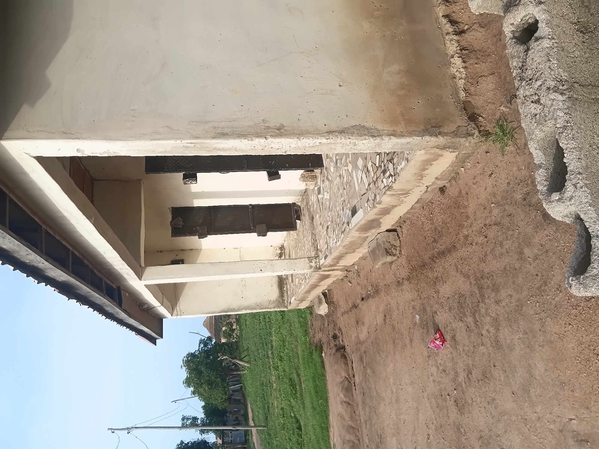 The image shows a simple, weathered building in Jalingo, Nigeria. The building has a covered porch with a tiled floor and several dark, metal-barred doors. The exterior walls are made of a rough, light-colored plaster that shows signs of age and some staining. In front of the building, the ground is a mix of dirt and some patchy grass. There is a piece of pink plastic litter on the ground. In the background, there are trees, some fencing, and a hut with a thatched roof, suggesting a rural or semi-rural setting within the city. The sky is clear and blue, indicating daytime. There are no people visible in the image.