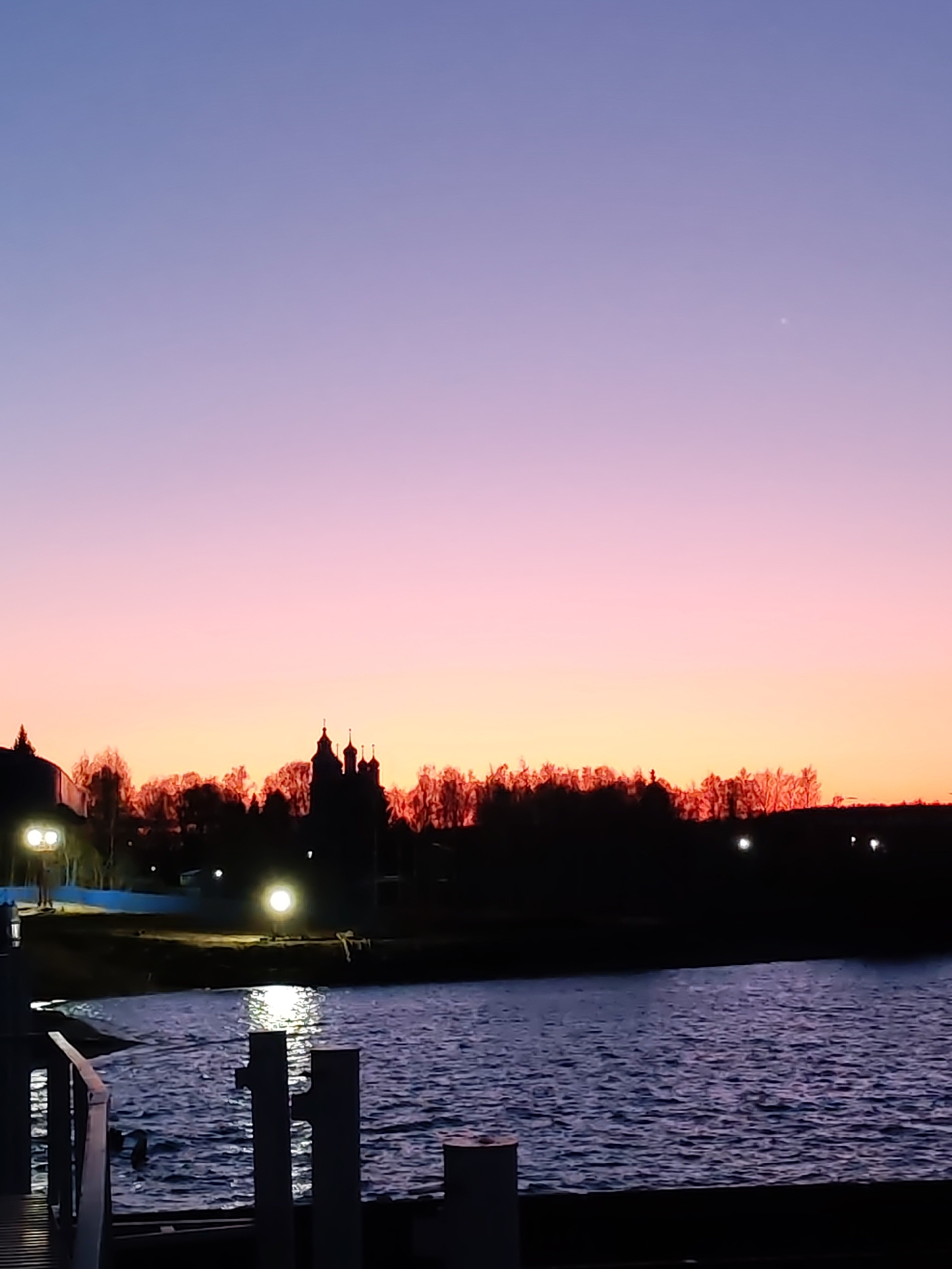 The image depicts a twilight scene over a body of water. In the foreground, there is a wooden dock or pier. The water reflects the lights on the shoreline. In the midground, there's a silhouette of a line of trees and buildings, including what appears to be a church with multiple domes. The background features a gradient sky transitioning from orange near the horizon to pink and purple above. A small, bright dot is visible in the upper right portion of the sky. The location is Unknown, Unknown.
