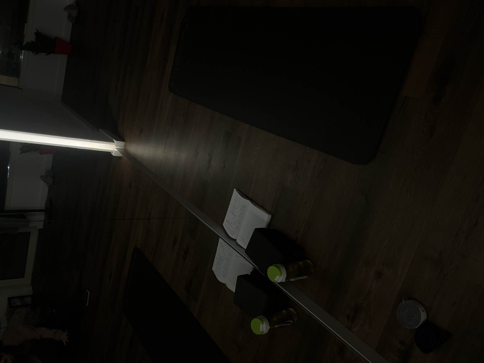 The image depicts an indoor scene with a dark, wooden floor. A long, rectangular mirror extends from the lower left to the upper right of the frame, reflecting the surroundings. Near the center of the image, a notebook is placed next to a black rectangular object. Two bottles with green lids are situated nearby. To the right of these items, there is a gray, rolled-up object. A dark yoga mat occupies the upper part of the frame. The location is not identifiable from the image.