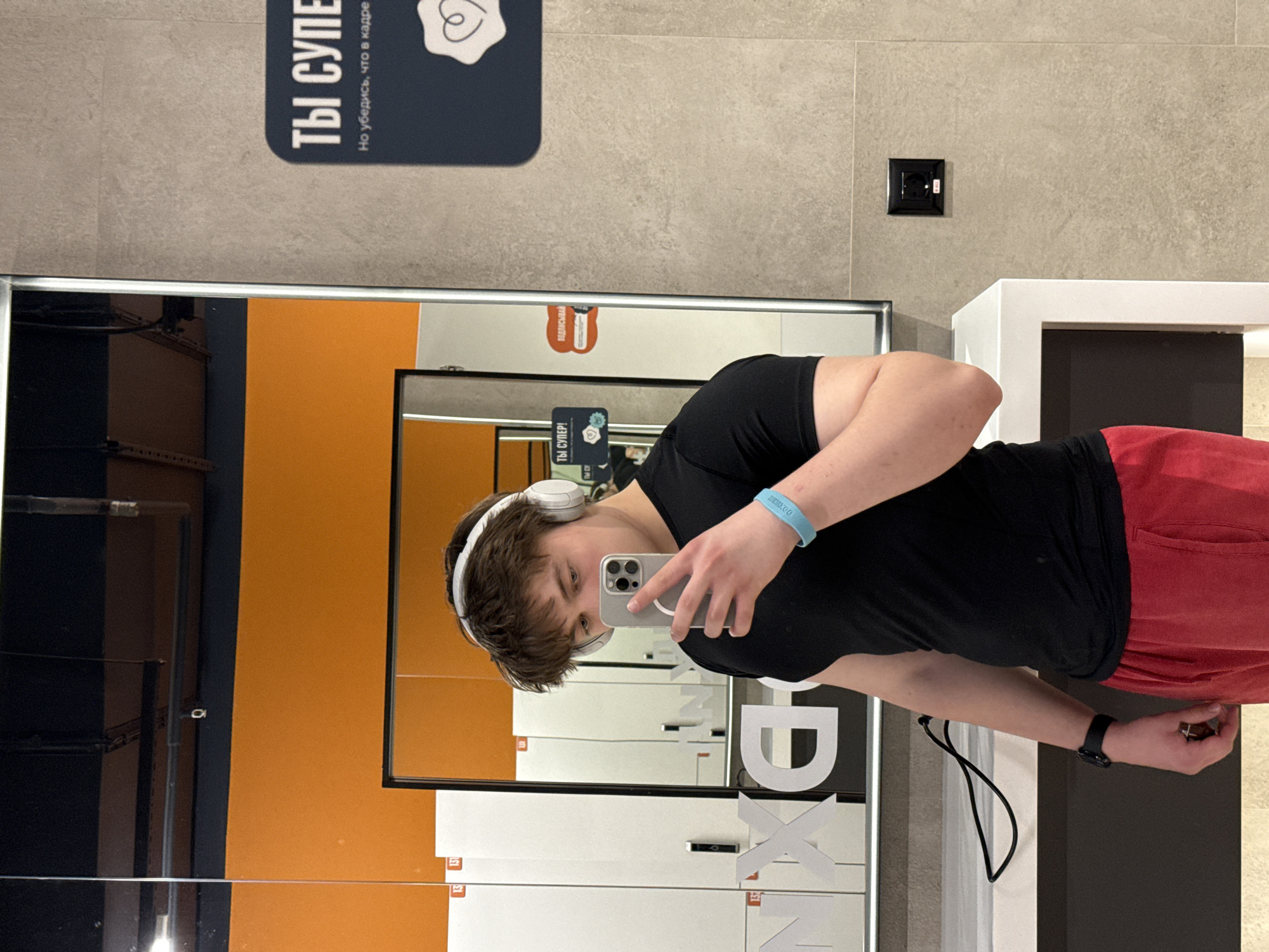 A young man is taking a selfie in a mirror at a location identified as the X-Fit gym in Krasnoyarsk, Russia. He is wearing a black T-shirt and red shorts, along with white headphones and a smart watch on his left wrist. He is holding a smartphone up to the mirror with his right hand. The background consists of lockers, an orange wall, and informational signs in Russian. The overall setting is a modern gym or fitness center.