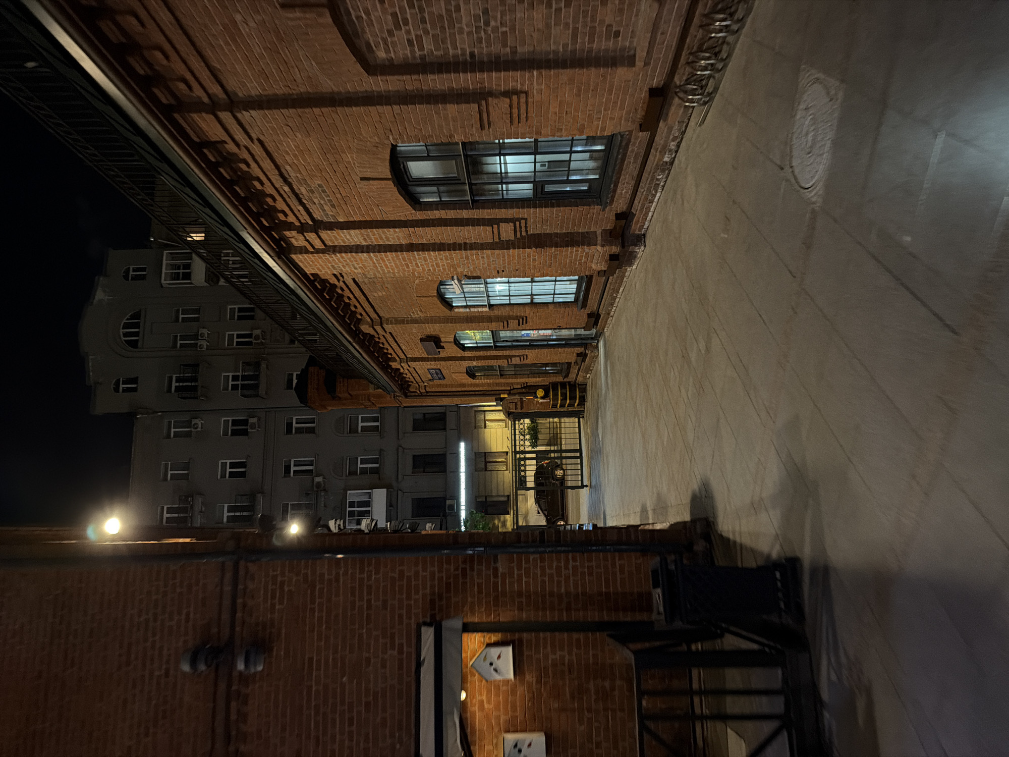 The image depicts an urban scene in Moscow, Russia, at night. The view is oriented along a narrow street or alleyway lined with brick buildings on either side. The building on the right features multiple windows. Across the alley is a taller building with numerous windows. Street lighting illuminates the buildings and pavement. A metal gate is visible at the end of the alley.
