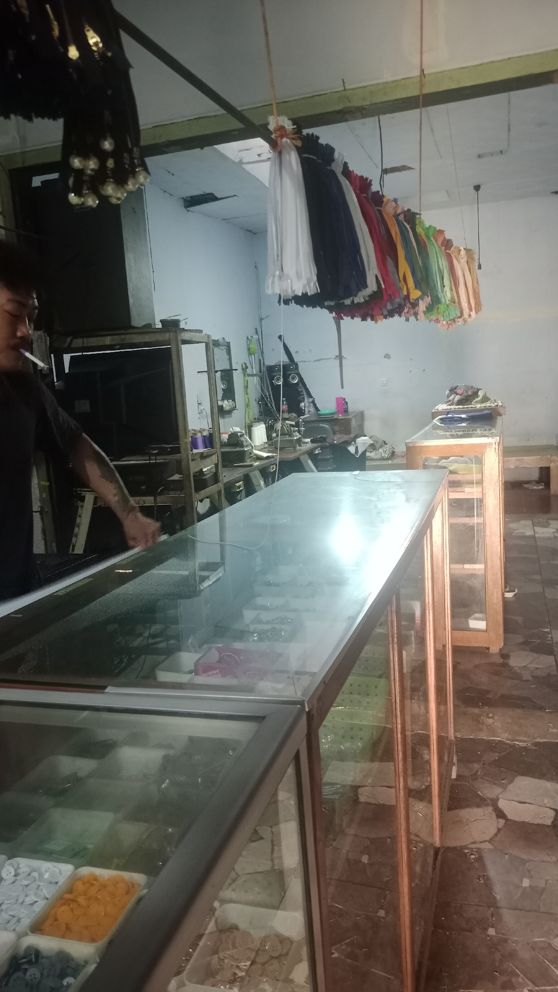 The image depicts an indoor market stall, likely in Pakemitan, Indonesia, filled with various items for sale. In the foreground, a glass-topped display case shows an assortment of buttons in different colors and sizes, arranged in plastic trays. To the left, a man wearing a black t-shirt is visible, smoking a cigarette and reaching towards the display case. Behind him, shelves are filled with more items. Hanging from the ceiling are numerous colorful fabrics or garments, creating a vibrant backdrop. Further into the stall, other display cases and shelving units hold more merchandise. The environment appears somewhat cluttered, suggesting a busy retail space. The flooring is tiled with a stone-like pattern, and the walls are painted a light color. The overall impression is of a small business selling sewing supplies or accessories.