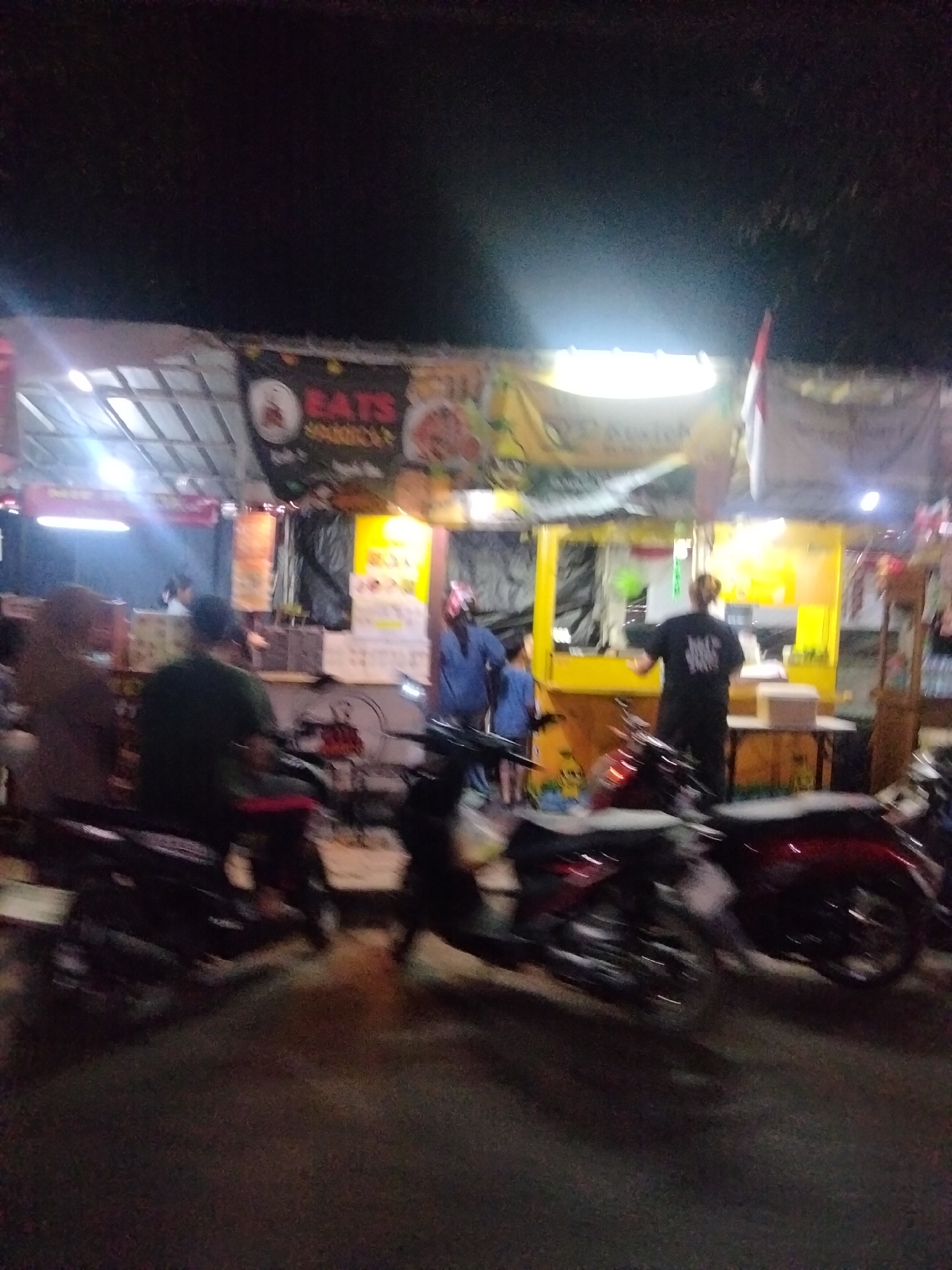 The image depicts a street scene at night, possibly a food stall area, in Cariu, Indonesia. A long, open-sided structure with a corrugated roof covers multiple food vendors. Bright fluorescent lights illuminate the scene. In the foreground, several motorcycles are parked, some appearing to be in motion due to motion blur. On the right side of the frame, a person in a black t-shirt stands behind a counter, facing away from the camera. To their left, a child in a blue shirt stands near another person. A banner with the word "EATS" in red lettering is visible above one of the stalls. The overall atmosphere suggests a bustling, informal commercial setting.