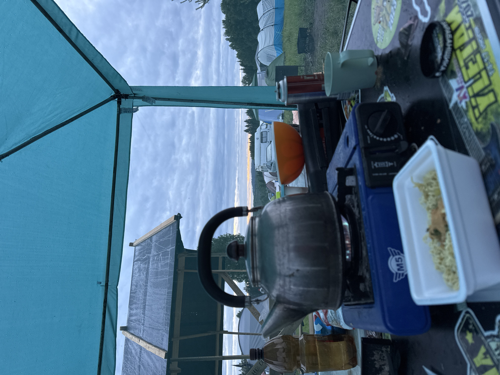 The image depicts an outdoor camping scene observed from underneath a teal-colored waterproof canopy, which is speckled with numerous water droplets. In the immediate foreground, a metallic kettle with a dark, arched handle is situated on a blue portable gas stove, emitting a visible flame. To the left of the stove, a brown plastic bottle is present. To the right, a white rectangular container holds dry instant noodles, and further right, a light green mug, a red aerosol can (likely butane for the stove), and a black circular object are visible on a dark-colored table. The table surface features reflective areas and various stickers, some with Cyrillic text.
Beyond the canopy, a wooden structure with a dark, potentially tarp-covered roof, also showing water droplets, stands to the left. The midground reveals a vast, grassy field dotted with numerous camping tents of varying sizes and colors, including a prominent white and blue striped tent on the right and a white tent with an orange band further left. A white van and a light-colored car are parked among the tents. The field extends to a dense forest line in the background. The sky is largely overcast with gray clouds, but a strip of lighter, warm-hued sky is visible near the horizon, indicative of either sunrise or sunset. No individuals are present in the frame. The scene is consistent with an outdoor gathering or camping event in a rural setting within the vicinity of Большая Кивара, Russia.