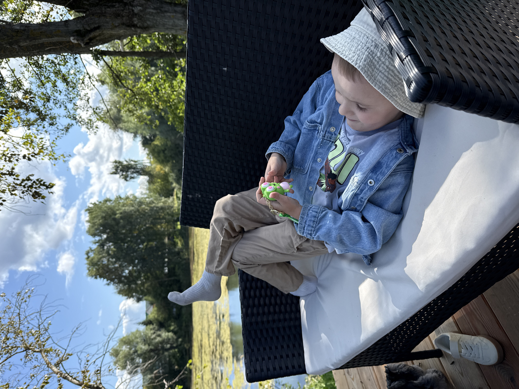 A young child is lying on a white cushioned outdoor sofa with a black woven frame, situated on a wooden deck. The child is wearing a light blue denim jacket over a t-shirt with a graphic print, tan pants, white socks, and a light-colored hat. One leg is bent with the foot extended upwards, and the other leg is bent at the knee. The child is holding a small green toy in their hands. The background shows a body of water covered in green aquatic plants, surrounded by trees under a bright blue sky with scattered clouds. To the left of the sofa, a white sneaker is visible on the wooden deck. In the far left foreground, a portion of a dark-furred animal, possibly a cat, is visible. The scene appears to be in Chaikovsky, Russia.