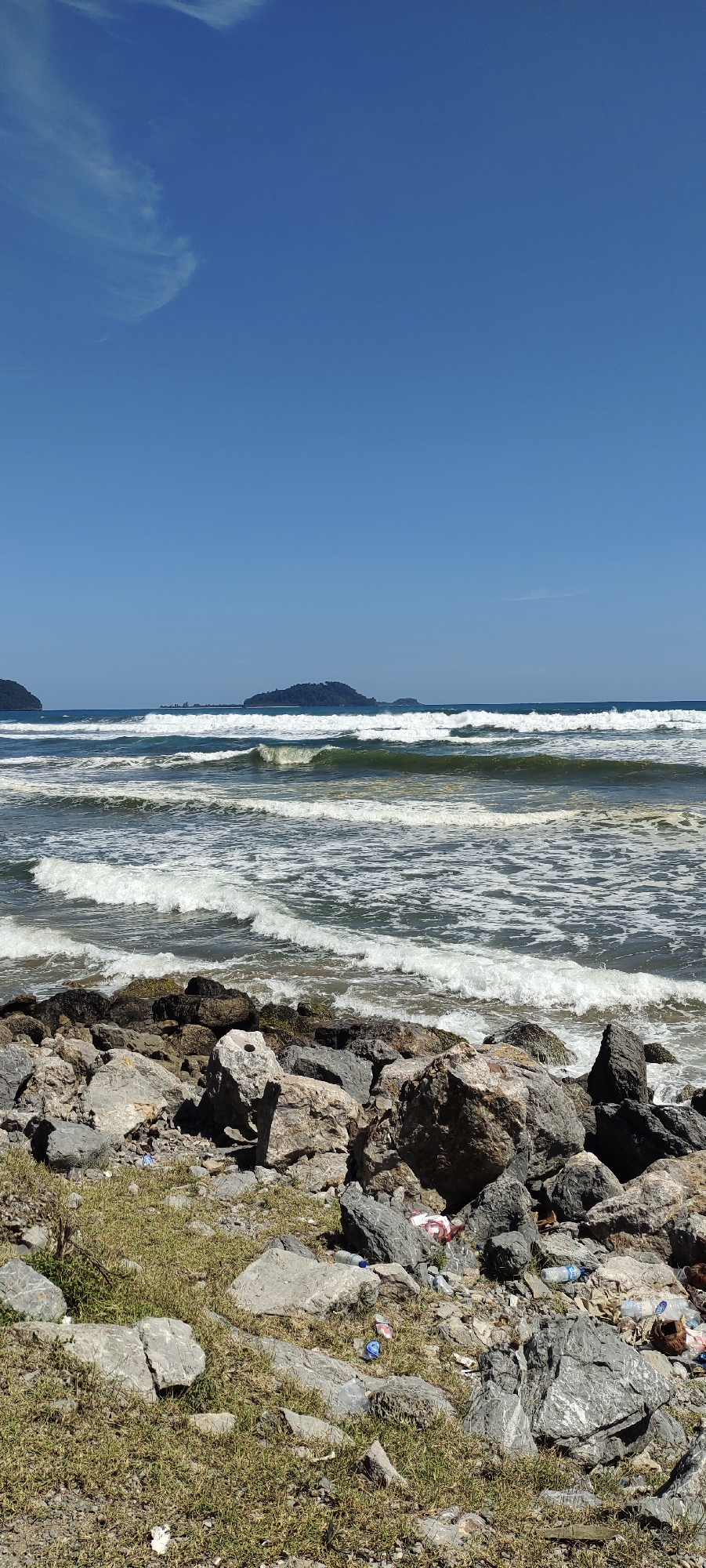The image depicts a coastal scene with a rocky foreground and the ocean stretching towards the horizon under a clear blue sky. Several islands are visible in the distance. The foreground is littered with various types of trash, including plastic bottles, wrappers, and other debris, scattered among rocks and patches of dry grass. The ocean exhibits moderate wave activity with white foam breaking along the shore. The overall impression is of a natural environment impacted by human waste. The location is identified as Meudheun, Indonesia.