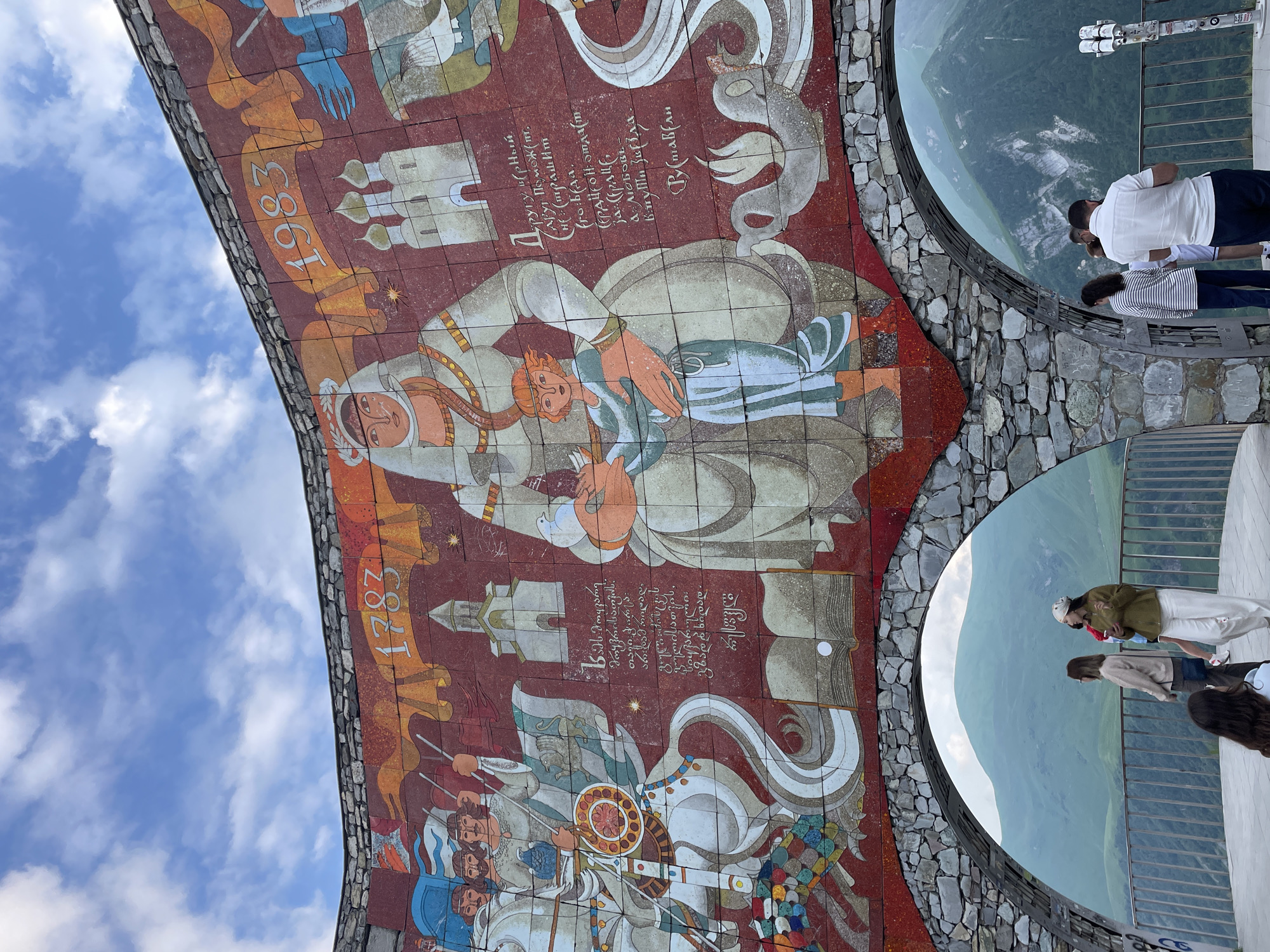 This is a medium shot, eye-level, outdoor photograph of the Gudauri Monument, also known as the Monument of the Russia–Georgia Friendship, located in Gudauri, Georgia. The monument is a large, circular mosaic mural on a cliffside, with arched openings offering views of the Caucasus Mountains.
The mural depicts historical scenes, with figures dressed in traditional attire, including soldiers on horseback, a central matriarchal figure holding a child, and architectural elements like churches and domes. There are inscriptions in both Georgian and Russian languages visible on the mural. The years "1783" and "1983" are prominently displayed. The mural is made of colorful tiles, and the sky above is blue with scattered white clouds.
In the foreground, under the monument's arches, several people are standing on a viewing platform. They are casually dressed and appear to be tourists observing the mountainous landscape. There are railings for safety. The environment is characterized by steep, green, and rocky mountain slopes under a cloudy sky. The overall scene conveys a sense of historical grandeur and natural beauty.