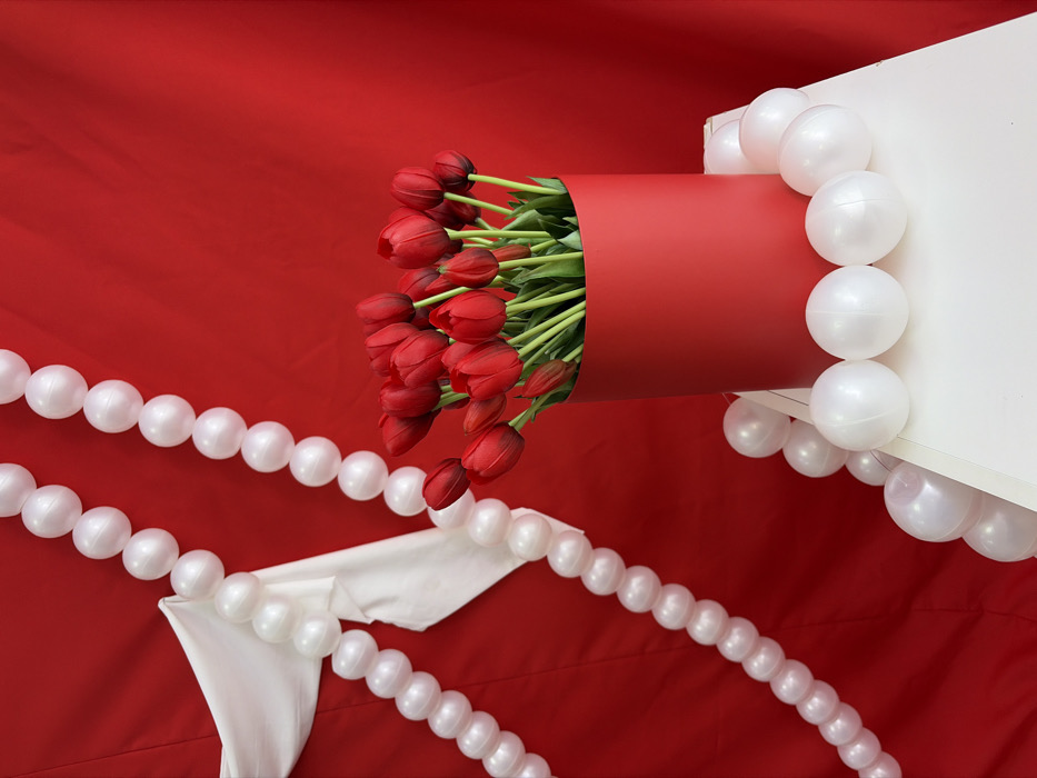The image displays an indoor decorative arrangement situated against a red fabric backdrop in Chaikovsky, Russia. A tall, cylindrical red container, approximately 1.5 times taller than its diameter, is centrally positioned. This container holds a dense bouquet of roughly 25-30 red tulips with prominent green stems. The tulips appear uniform, suggesting artificiality. The red container rests upon a white, multi-faceted pedestal or display stand. Two vertical strands of white, spherical, pearl-like objects are visible in the upper left and mid-left areas, descending diagonally. A third strand of these white spheres is arranged horizontally around the base of the red container on the white pedestal. A piece of white fabric is draped and folded over one of the vertical strands of white spheres in the upper left quadrant of the frame. The red backdrop exhibits minor wrinkles and folds. No individuals are present in the scene.