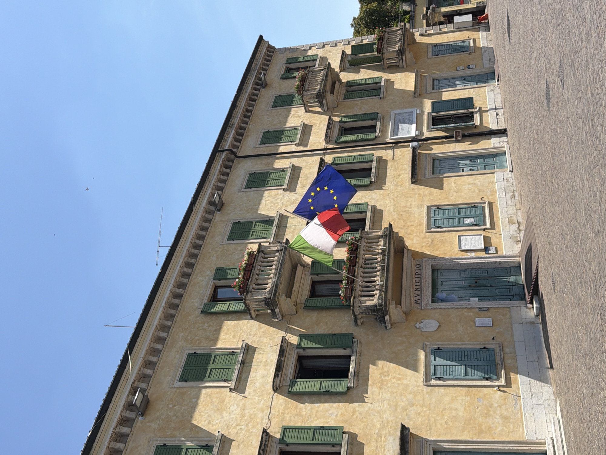 The image shows a building facade with yellow stucco and green shutters on a sunny day. The building appears to be the Town Hall (Municipio) of Peschiera del Garda, Italy, as indicated by the inscription above the central entrance. Two flags are prominently displayed on balconies: the Italian flag and the flag of the European Union. Several windows, some open and some closed, are visible. Flower boxes with red and white flowers adorn the balconies. The building's entrance is a double-door painted teal, with a ramp leading up to it. The ground in front of the building is paved with cobblestones. In the background, a tree-lined area and parts of other buildings are visible. The sky is clear and blue. There are no people visible in the scene.