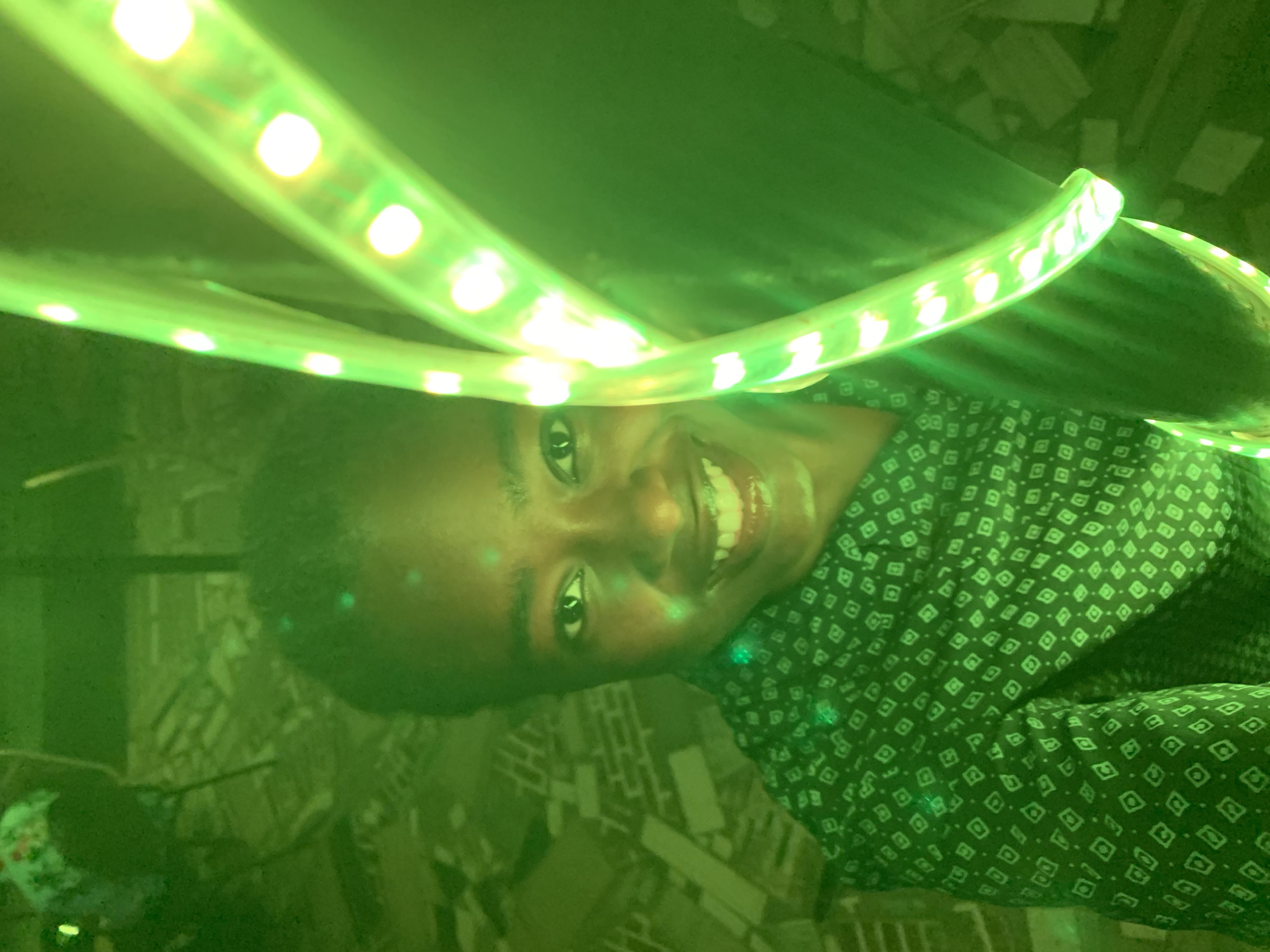 A close-up, tilted shot features a person with dark skin and short dark hair smiling broadly. The person is wearing a dark shirt with a diamond pattern. Bright green LED strip lights curve around their head, casting a green glow on their face and clothing. The background appears to be an indoor setting with indistinct features. There is no confirmation that the location is New Stadium, Nigeria.