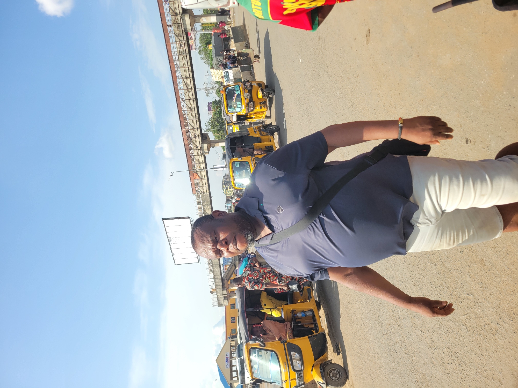 This image, presented at a 90-degree counter-clockwise rotation, captures a man standing on a bustling street in Fegge, Nigeria, under a bright, clear blue sky.
The main subject is a dark-skinned man with a beard and short hair, wearing a dark blue polo shirt and light-colored trousers. A black bag strap crosses his chest, and he has a bracelet on his left wrist. He is looking towards the camera with a slight smile, appearing calm.
The background reveals an active urban environment. Several yellow tricycles, locally known as Keke Napep, are prominent on the road, with some carrying passengers. One tricycle has the text "KINGSH" visible on its front. An elevated concrete and metal structure, possibly a bridge or overpass, spans across the middle ground. Further in the distance, other vehicles, buildings, and a large, empty billboard frame can be seen.
The bright sunlight and distinct shadows indicate that it is daytime, likely mid-morning or early afternoon, with clear weather and a few scattered white clouds. The scene portrays a typical moment of daily life and urban transportation in the region. A fragment of text, "ORTU", is partially visible on a red and green fabric in the upper right corner of the overall image.