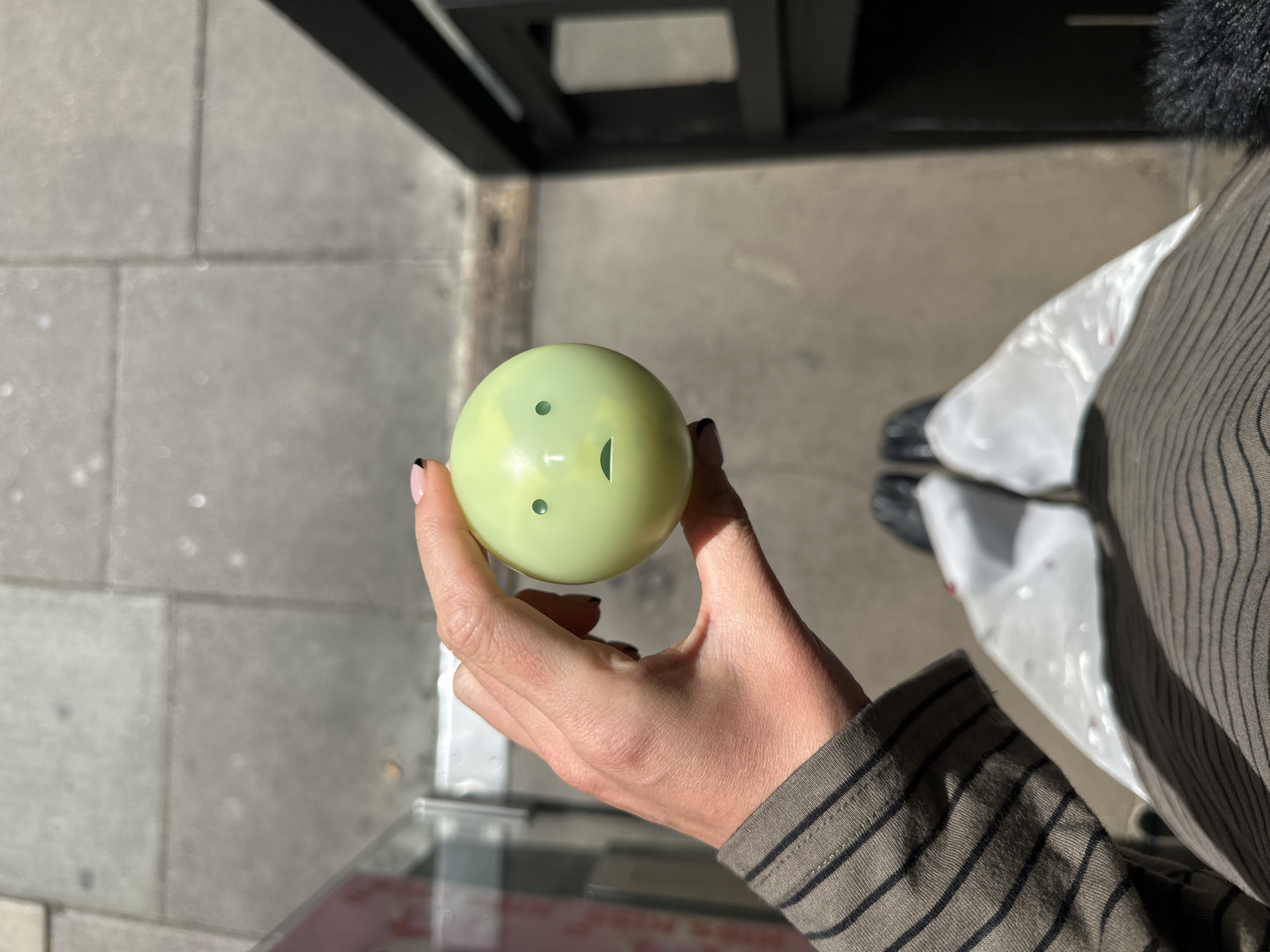 A person holds a light green gumball machine capsule in their right hand. The capsule has two round holes and a slit cut in the center, resembling a face. The person is wearing a striped shirt, and their fingers are visible, with black nail polish. The location appears to be a city street, possibly in London, United Kingdom, based on the architectural details in the background: a black metal structure, possibly part of a building or storefront, and paving stones on the ground. A plastic bag and shoes are visible in the background as well.