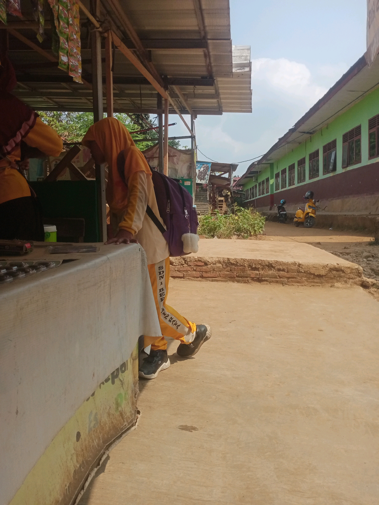 An outdoor, eye-level, medium shot shows a person wearing an orange hijab and orange pants with white lettering on the side, "SDN 1 BER..." and a purple backpack with white fuzz on the strap. The person is standing next to a counter. In the background, there is a green building with brown trim and many windows. Several yellow motorbikes are parked in front of the building. The sky is blue with a few white clouds. The ground is made of concrete. The scene is likely a school or a street near a school in Bandar Lampung, Indonesia.