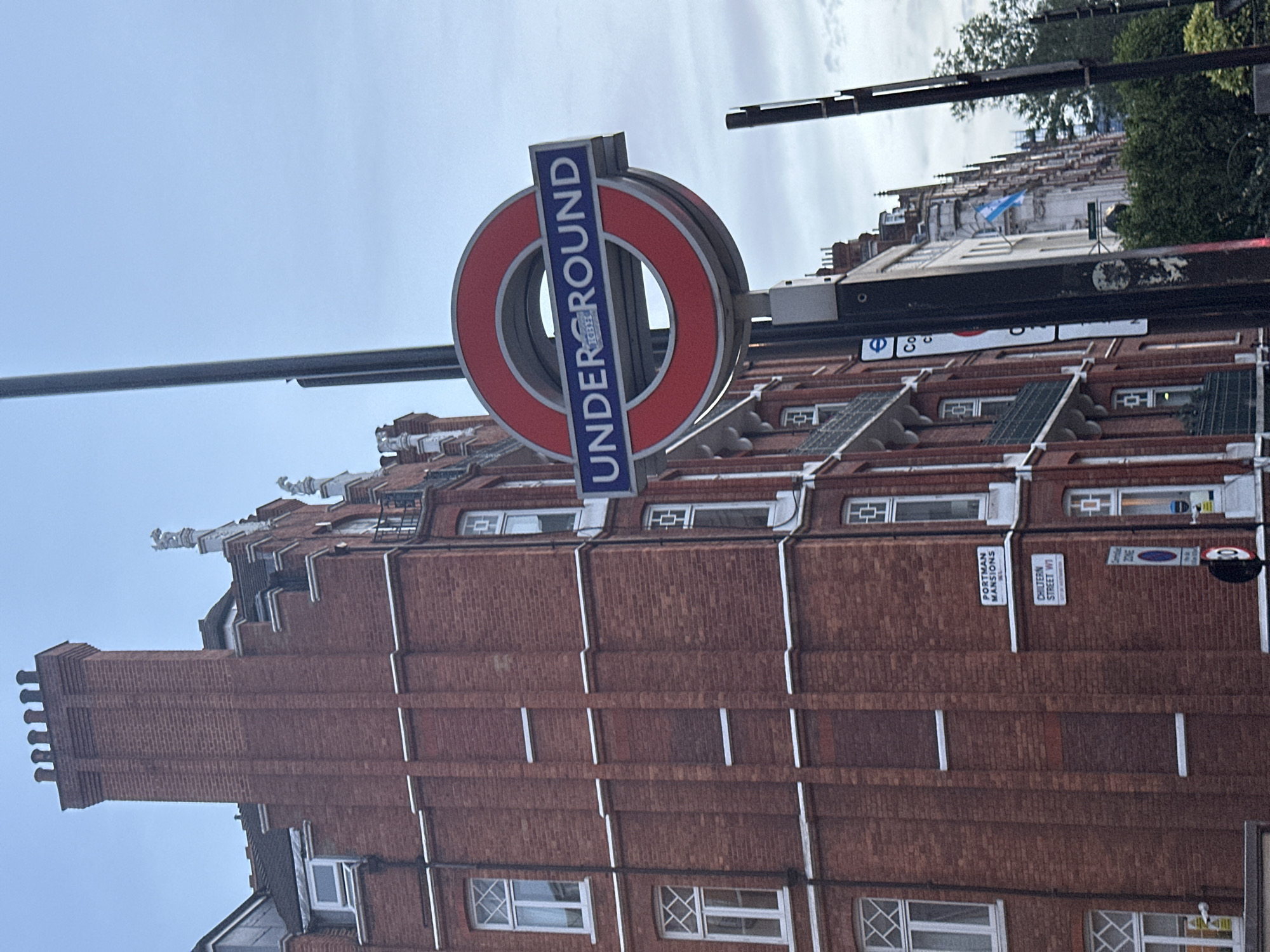 The image features a London Underground sign in front of a red brick building. The sign displays the circular logo of the Underground with the word "UNDERGROUND" written vertically. The building has a decorative facade with windows. A street sign for "Chiltern Street W1" and "Portman Mansions" is also visible.