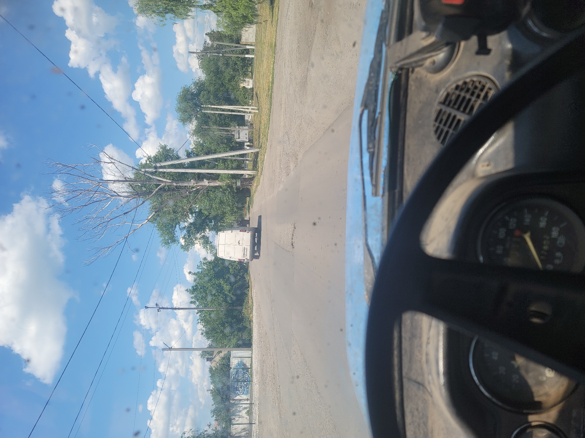 The image is a first-person perspective from inside a vehicle, looking out through the windshield. A steering wheel is visible in the foreground. The vehicle is on a paved road in what appears to be a rural or suburban area. A white van is driving away from the camera in the middle of the road. There are trees and buildings lining the road. Telephone poles and power lines are also visible. The sky is blue with white clouds. The scene is likely Pereshchepyne, Ukraine, given the context.