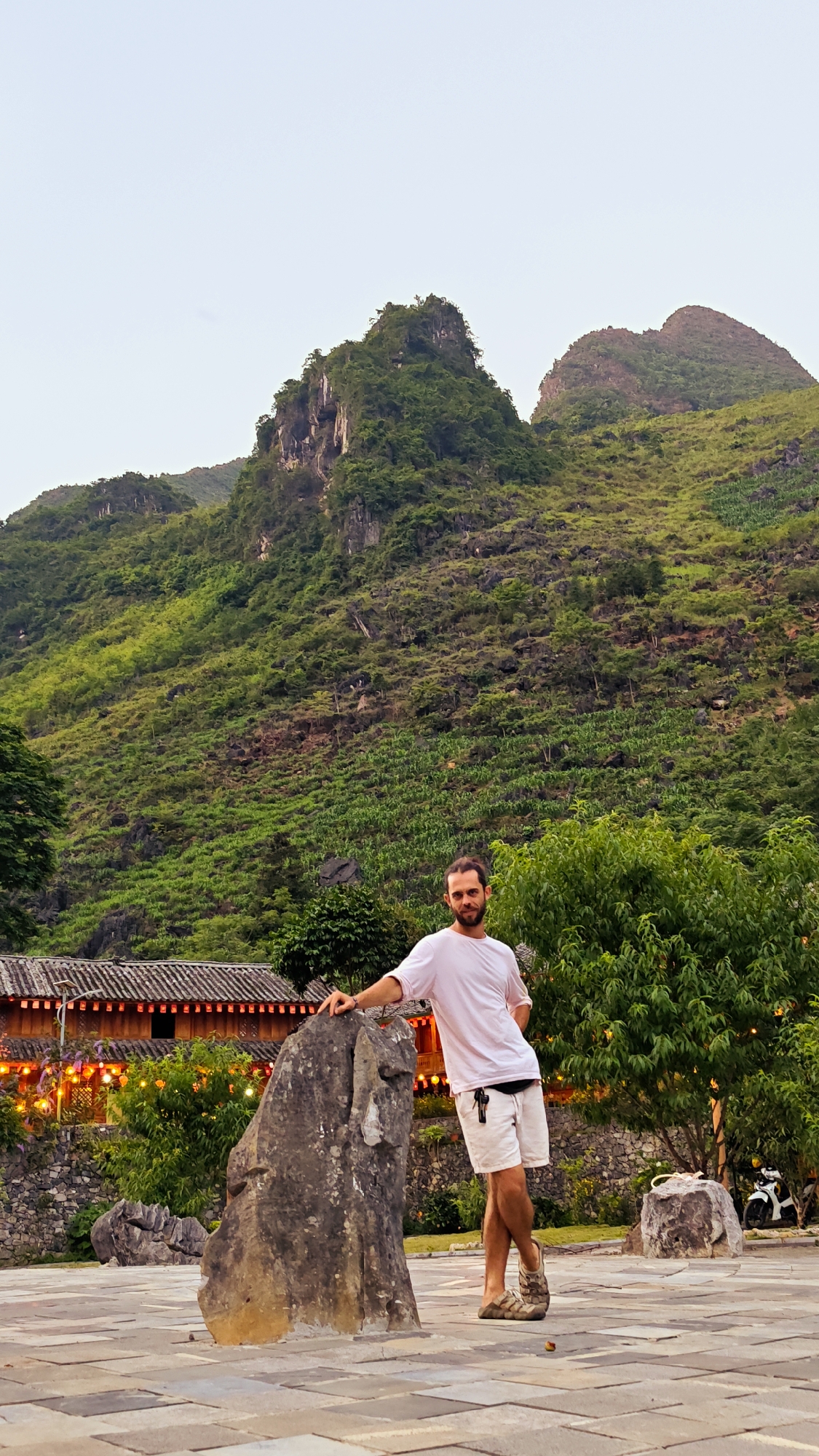 A full shot captures a man leaning on a rock in Mèo Vạc Commune, Vietnam. The man has a light beard, wears a white t-shirt, white shorts, and brown sandals. He leans casually with one arm on the rock, and his legs crossed at the ankles. Behind him is a building with a tiled roof decorated with string lights. In the background, two mountains covered in green vegetation rise up. The ground is made of light brown stone tiles. Other greenery, including trees and bushes, fills the space around the man and the building.