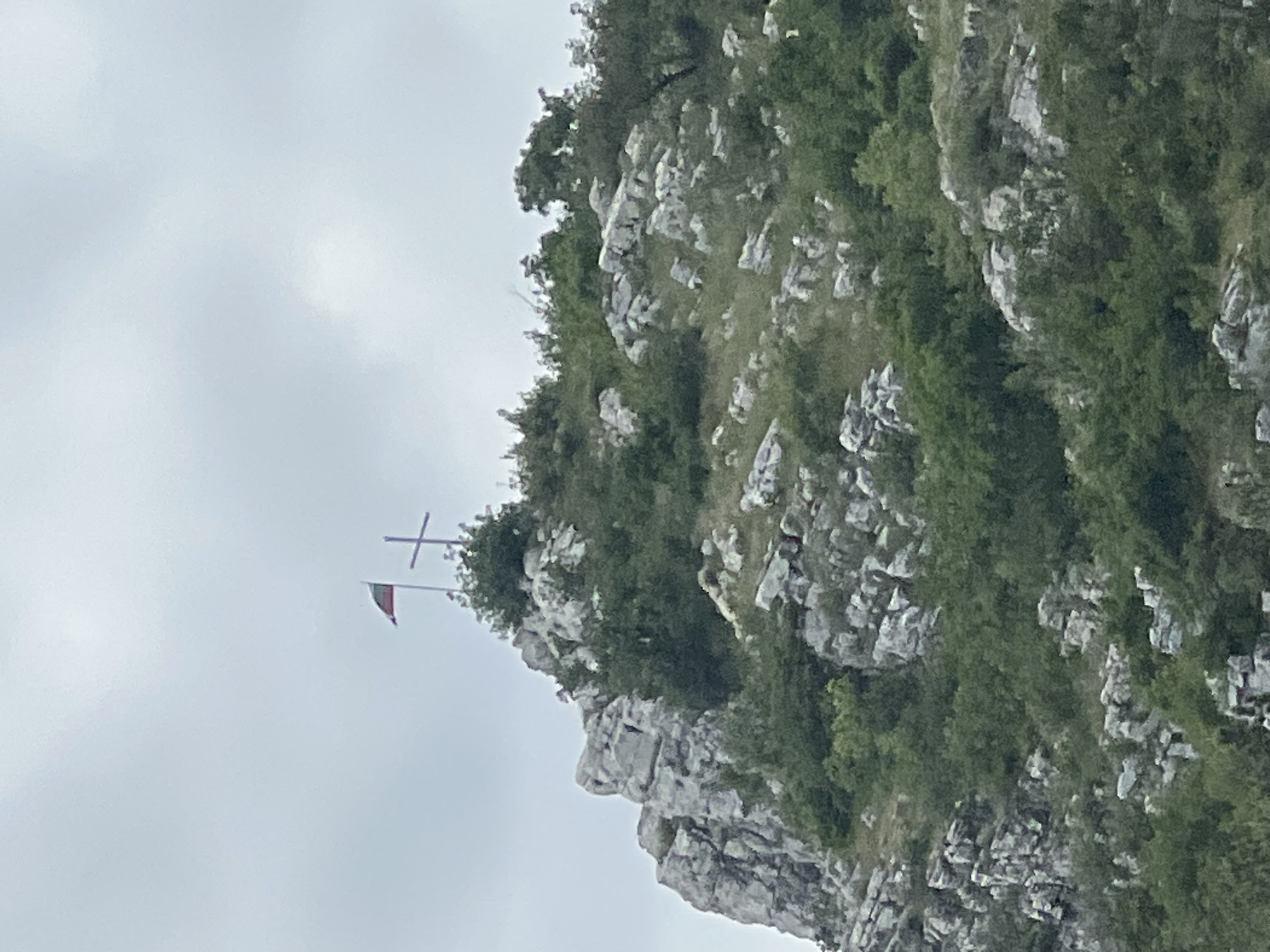 The image shows a rocky mountain slope with vegetation, a cross, and a flag on top. The flag appears to be red and white horizontally striped. The scene is taken in Asenovgrad, Bulgaria.